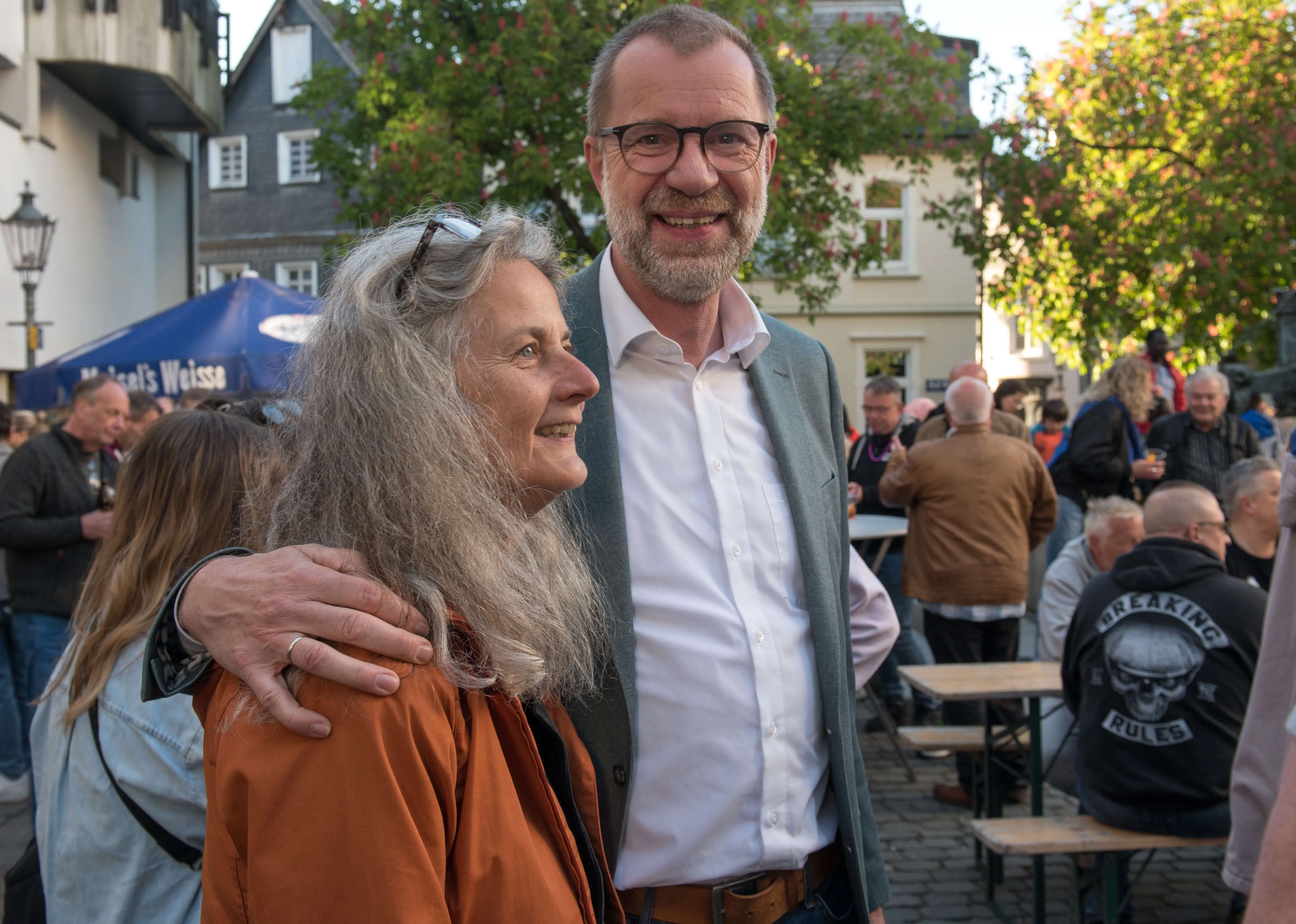 Ein lächelnder Mann mit Brille hat seinen Arm um die Schulter einer älteren Frau gelegt. Im Hintergrund sind Menschen auf einer belebten Straße zu sehen.