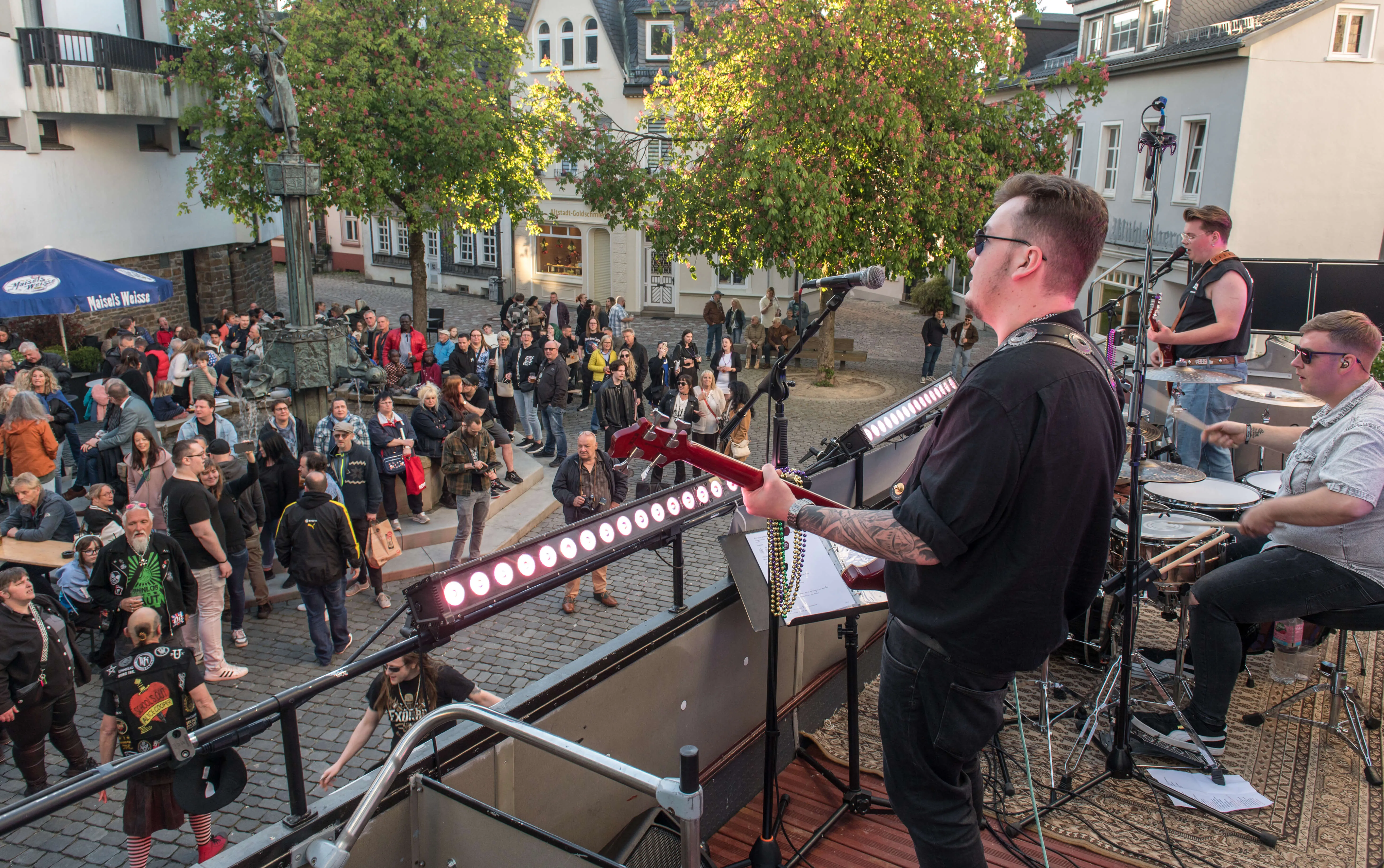 Ein Musiker spielt Gitarre auf einer kleinen Bühne vor einem großen Publikum auf einem Stadtplatz. Im Hintergrund sind Gebäude und weitere Menschen zu sehen.