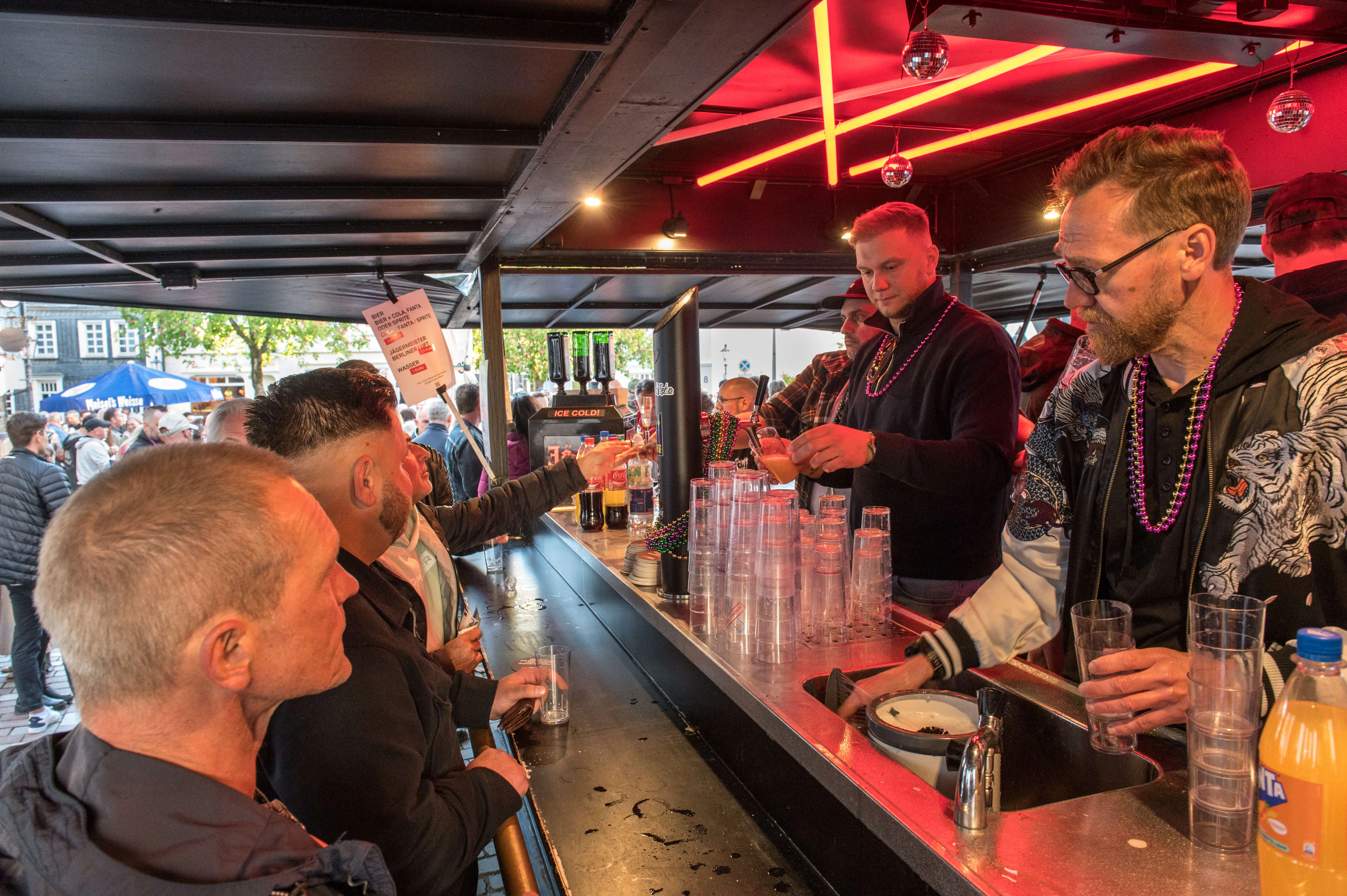 Ein Mann mit Bart und Perlenkette zapft an einer Bar ein Getränk für einen Gast. Im Hintergrund sind weitere Menschen und beleuchtete Theken zu sehen.