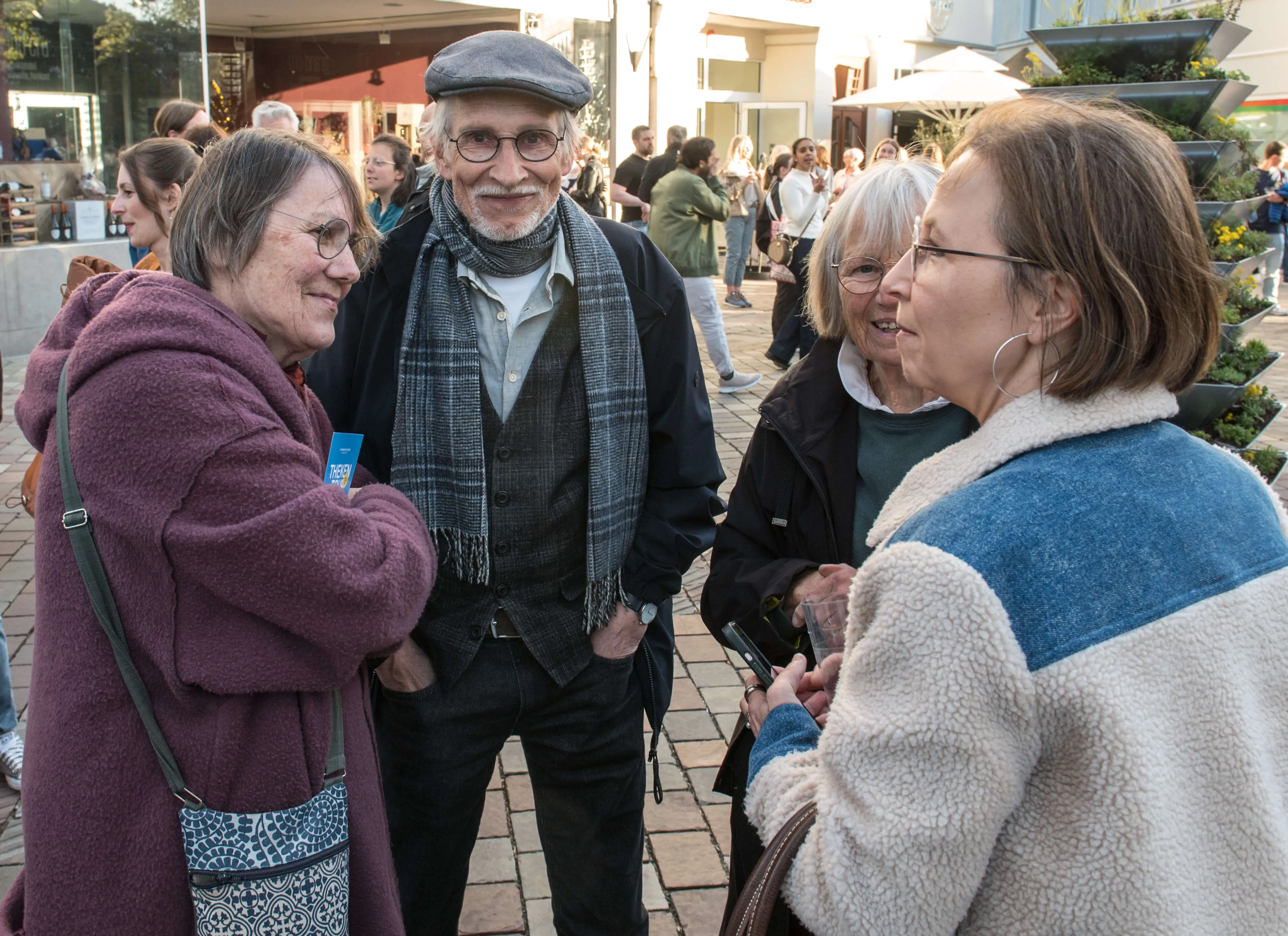 Drei ältere Menschen stehen im Gespräch auf einem belebten Platz, im Hintergrund weitere Passanten und Geschäfte.