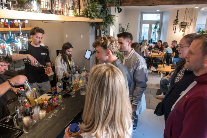 Eine Gruppe junger Erwachsener steht an einer Bar, während ein Barkeeper Getränke ausschenkt. Im Hintergrund sind weitere Gäste und eine geschäftige Atmosphäre zu sehen.