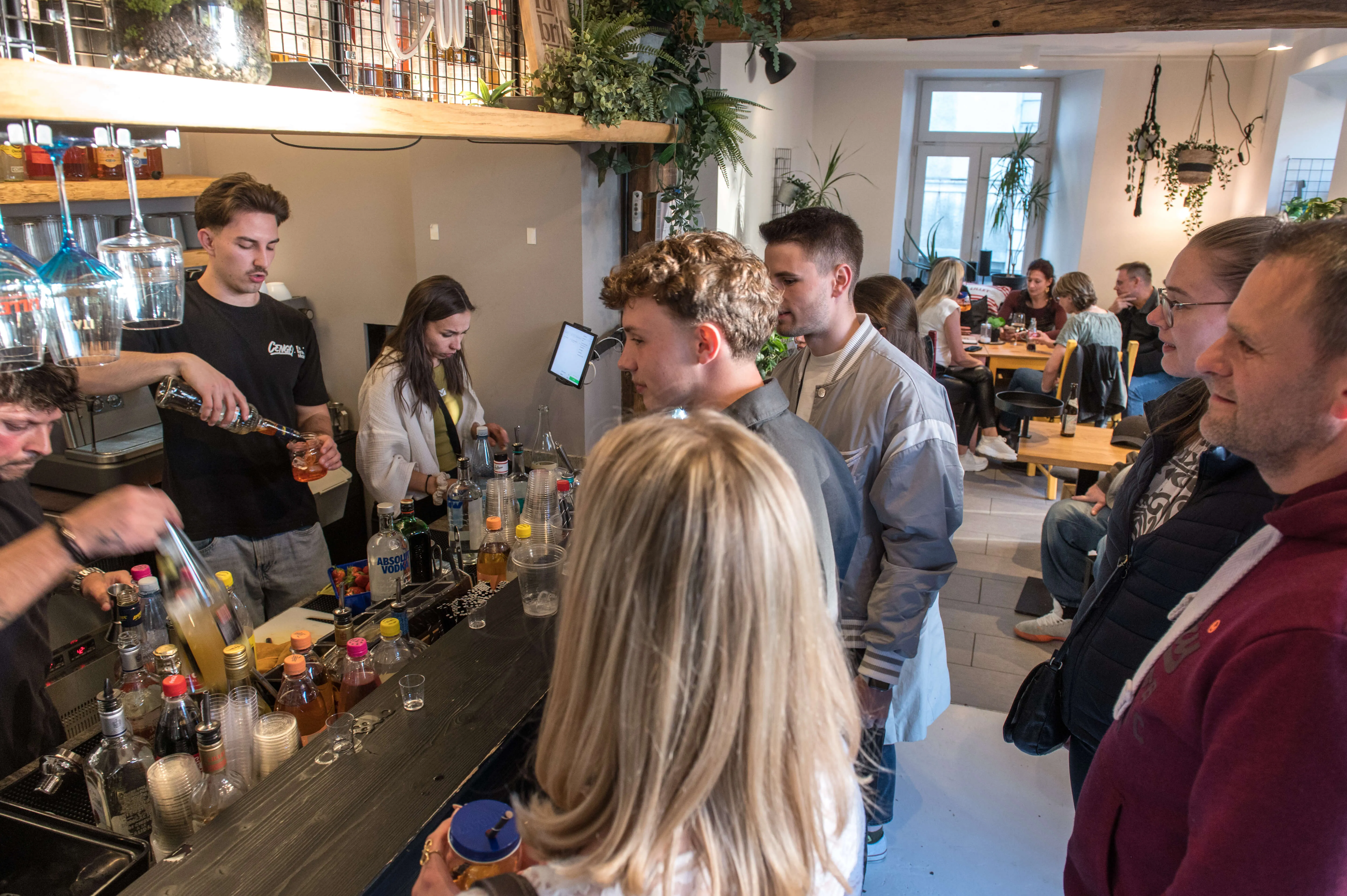 Eine Gruppe junger Erwachsener steht an einer Bar, während ein Barkeeper Getränke ausschenkt. Im Hintergrund sind weitere Gäste und eine geschäftige Atmosphäre zu sehen.