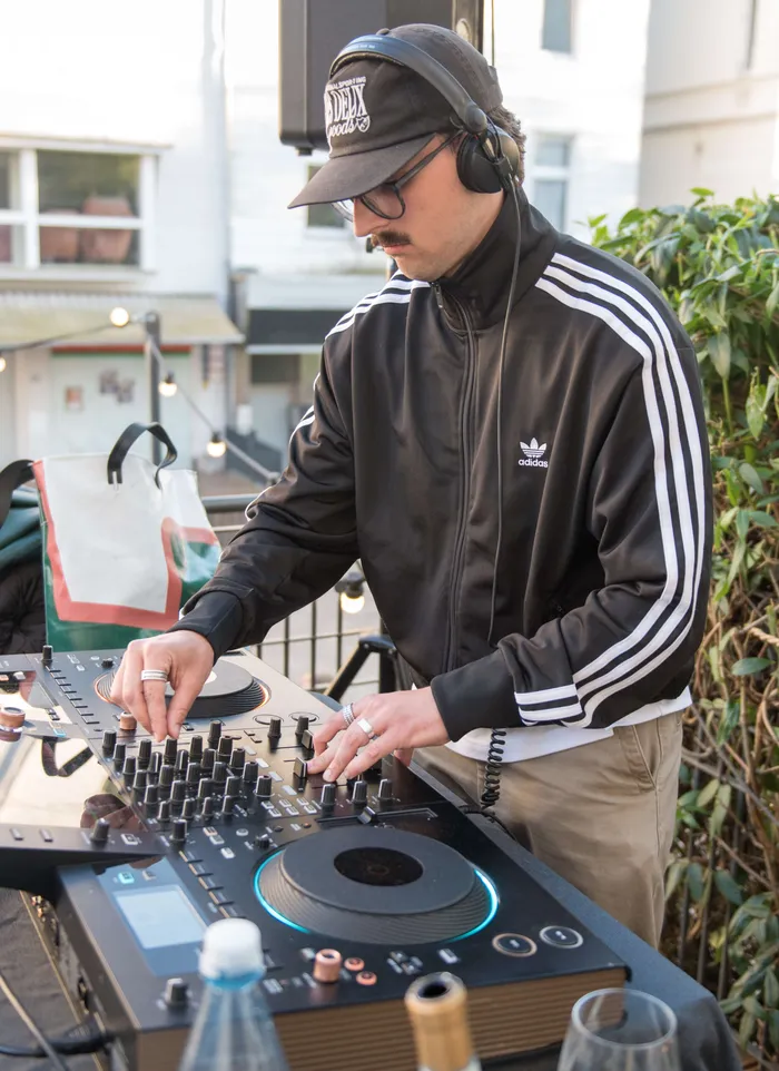Ein Mann mit Kopfhörern bedient ein DJ-Pult vor einer urbanen Kulisse mit Gebäuden und Büschen.