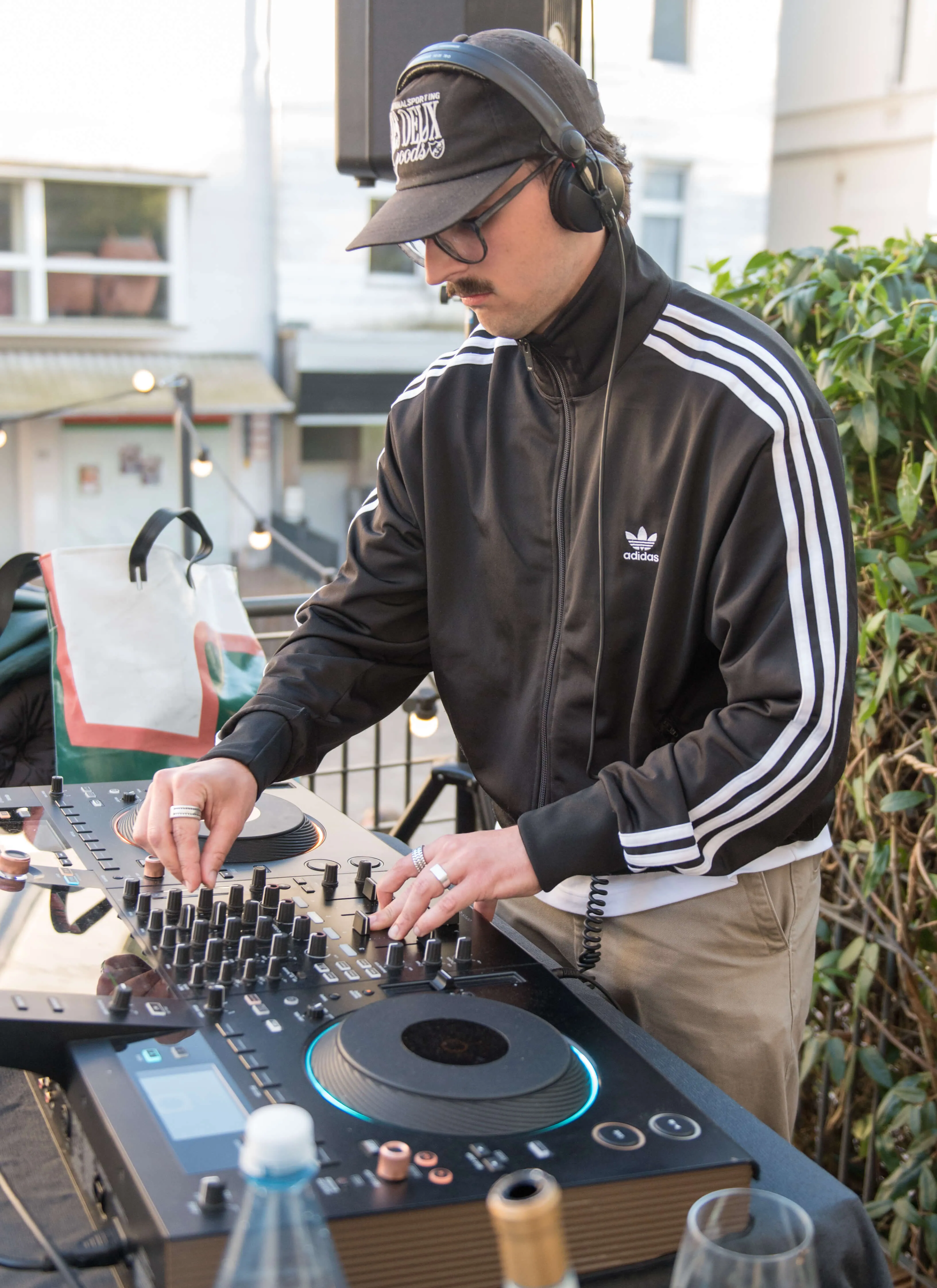 Ein Mann mit Kopfhörern bedient ein DJ-Pult vor einer urbanen Kulisse mit Gebäuden und Büschen.