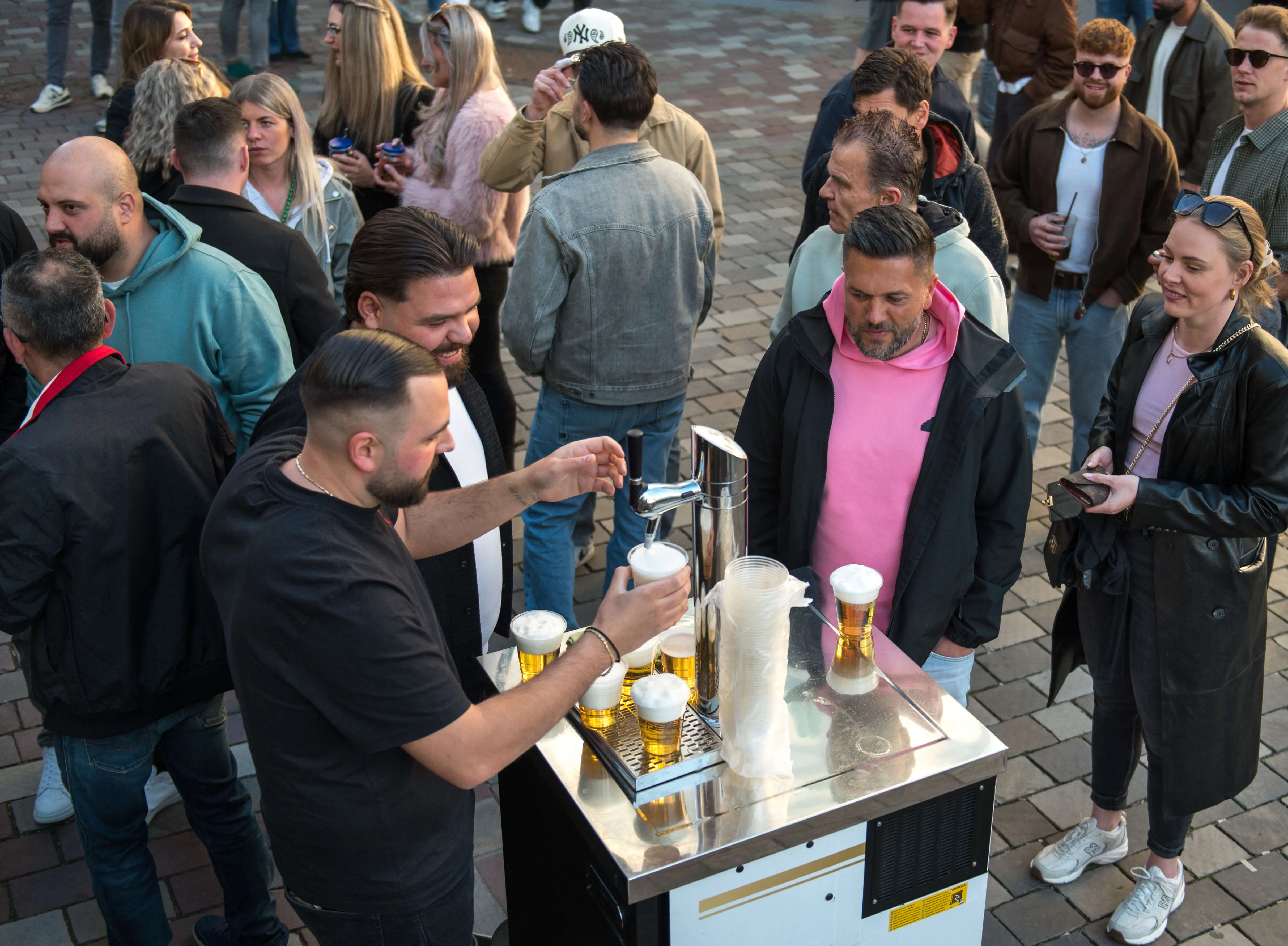 Ein Mann zapft Bier an einem mobilen Ausschankwagen, während sich um ihn herum eine Menschenmenge versammelt und Getränke genießt. Im Hintergrund erstreckt sich eine belebte Fußgängerzone mit weiteren Besuchern und Geschäften.