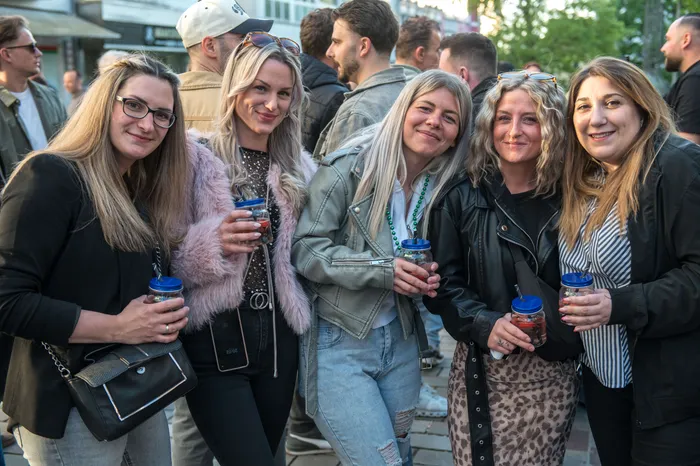 Fünf lächelnde Frauen mit Getränken in der Hand stehen inmitten einer Menschenmenge auf einer belebten Straße.