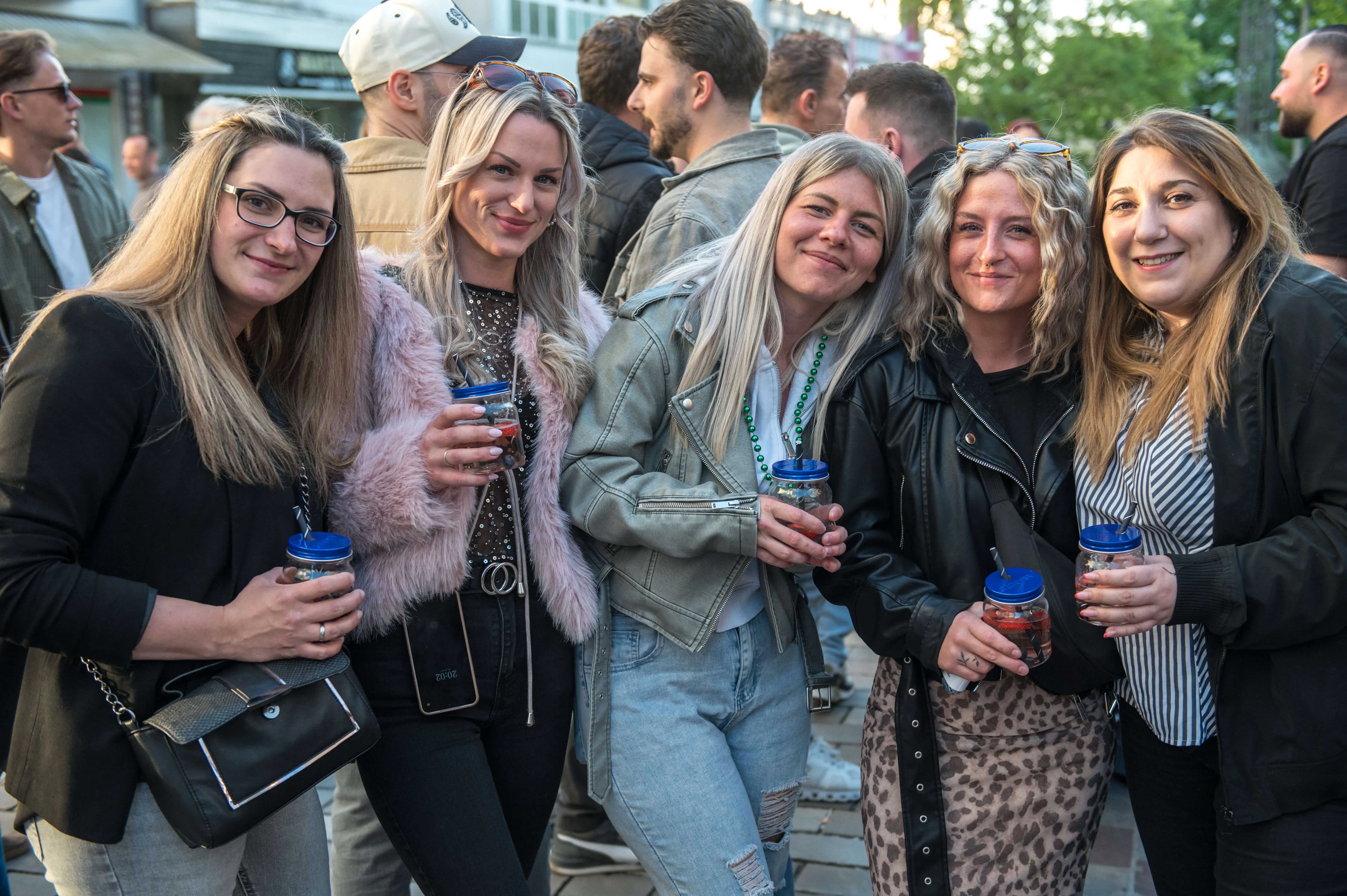 Fünf lächelnde Frauen mit Getränken in der Hand stehen inmitten einer Menschenmenge auf einer belebten Straße.