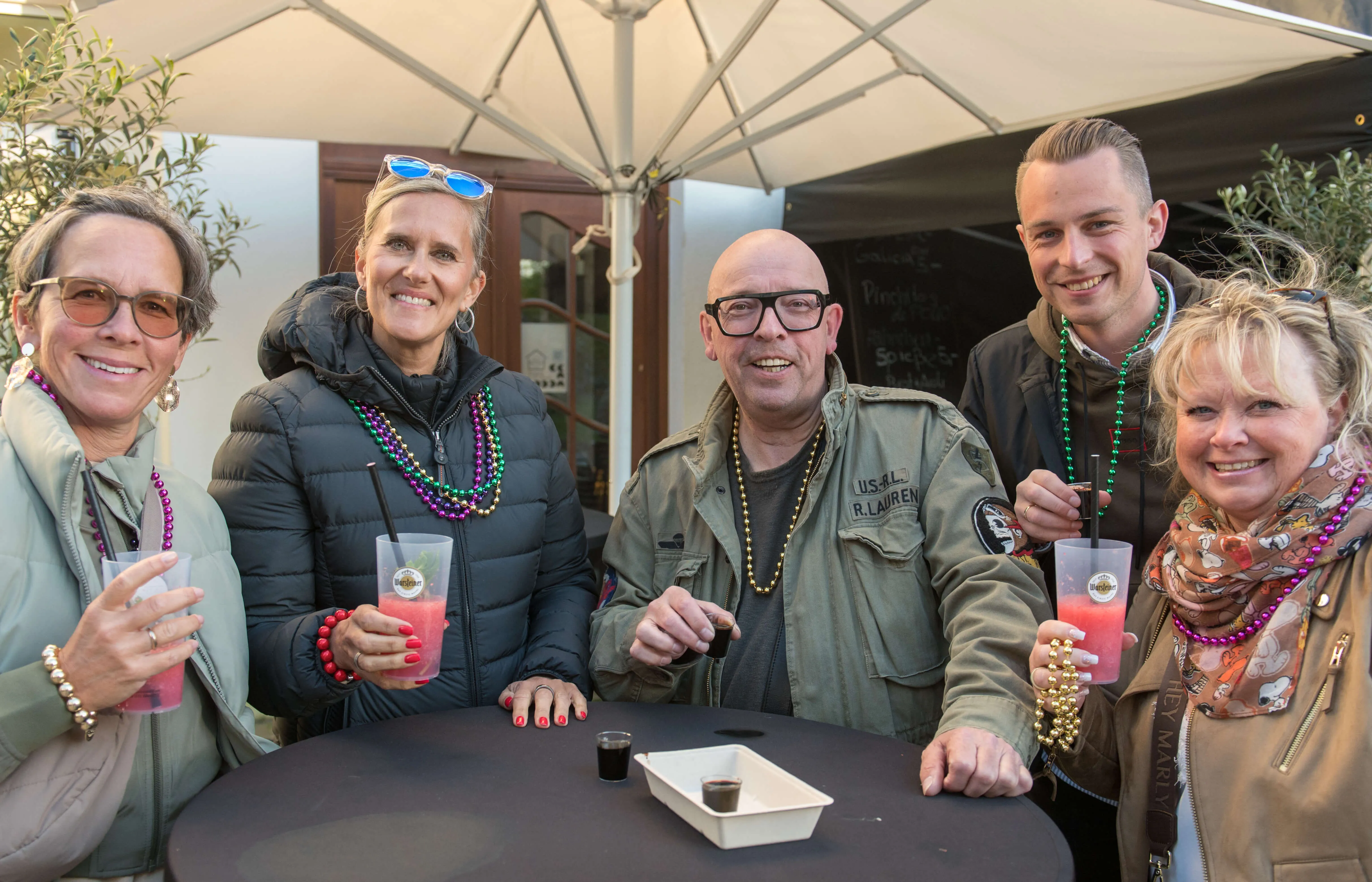 Fünf lächelnde Menschen sitzen an einem Tisch im Freien und halten bunte Getränke. Sie tragen Karnevalsketten und genießen die Atmosphäre der Thekentour in Lüdenscheid.