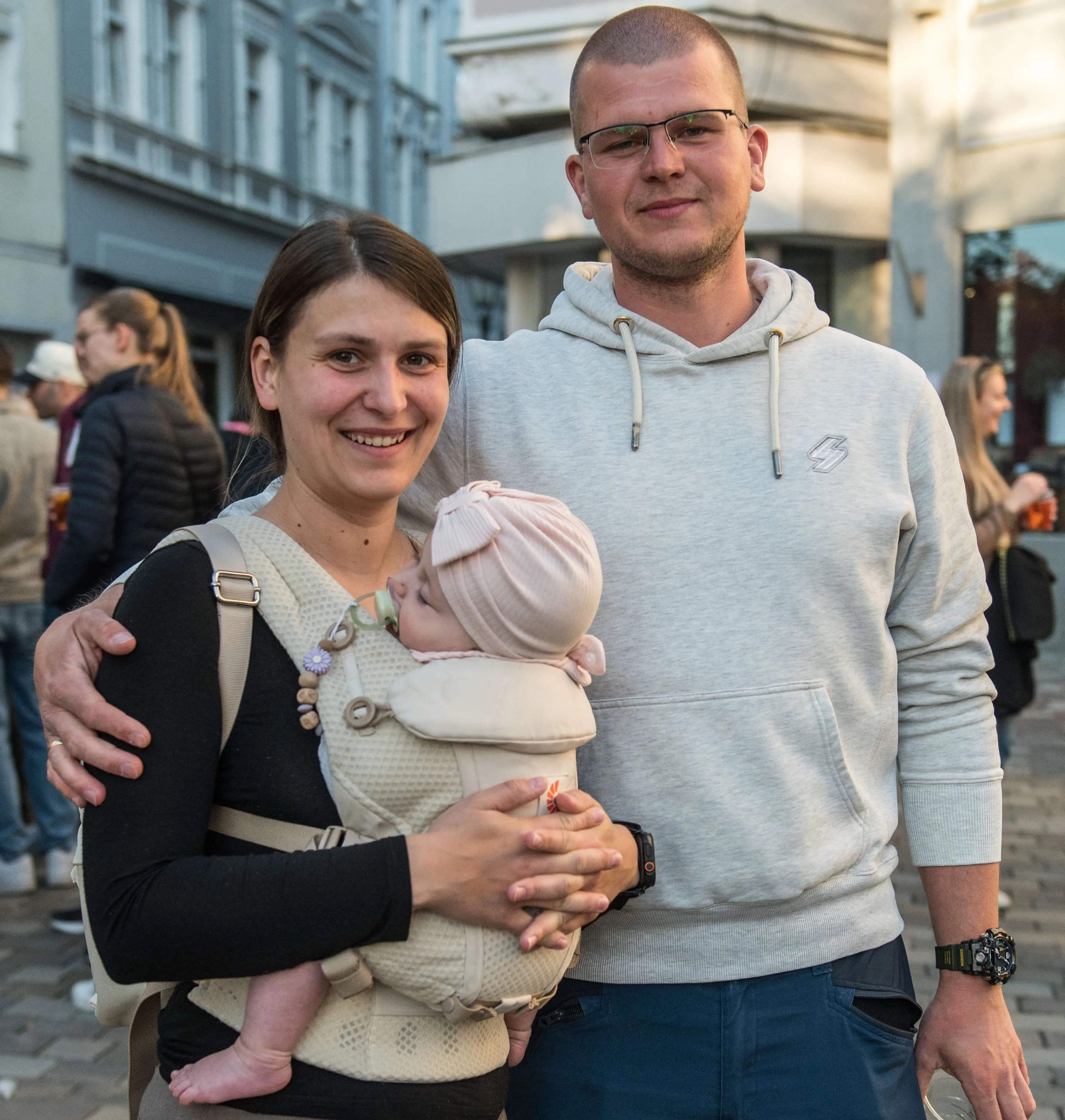 Ein Paar lächelt in die Kamera, die Frau hält ihr Baby im Tragetuch. Im Hintergrund sind weitere Menschen und Gebäude zu sehen.