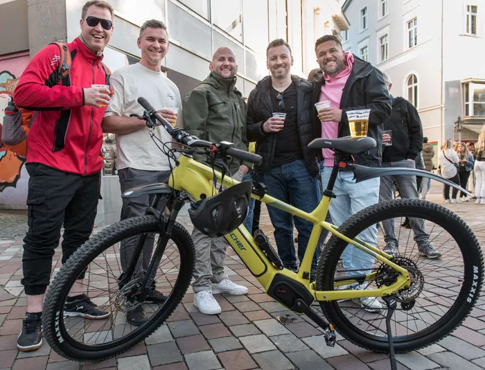Fünf Männer stehen lächelnd um ein gelbes E-Bike herum und halten jeweils ein Glas Bier in der Hand. Im Hintergrund ist eine belebte Fußgängerzone mit weiteren Menschen zu sehen.
