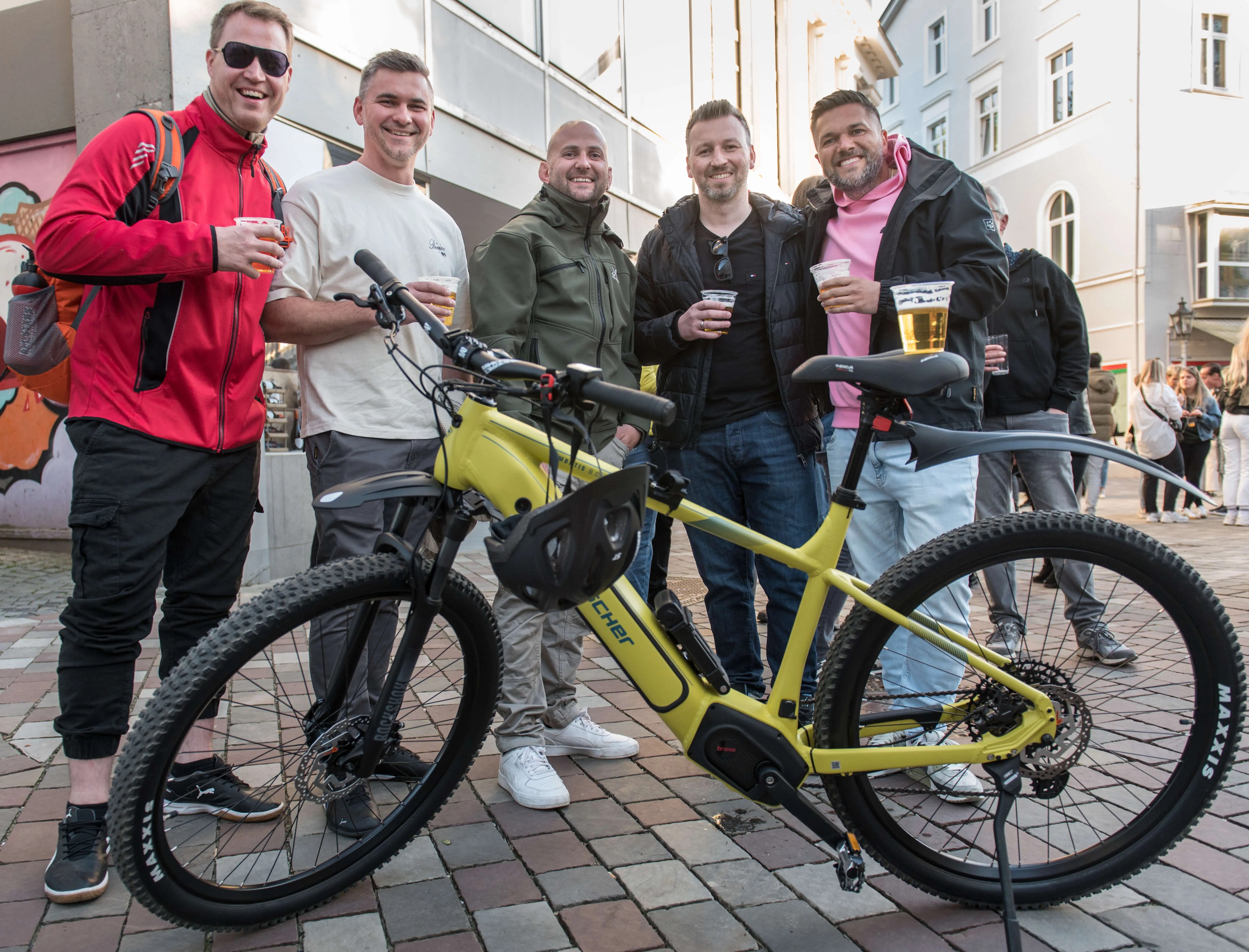 Fünf Männer stehen lächelnd um ein gelbes E-Bike herum und halten jeweils ein Glas Bier in der Hand. Im Hintergrund ist eine belebte Fußgängerzone mit weiteren Menschen zu sehen.