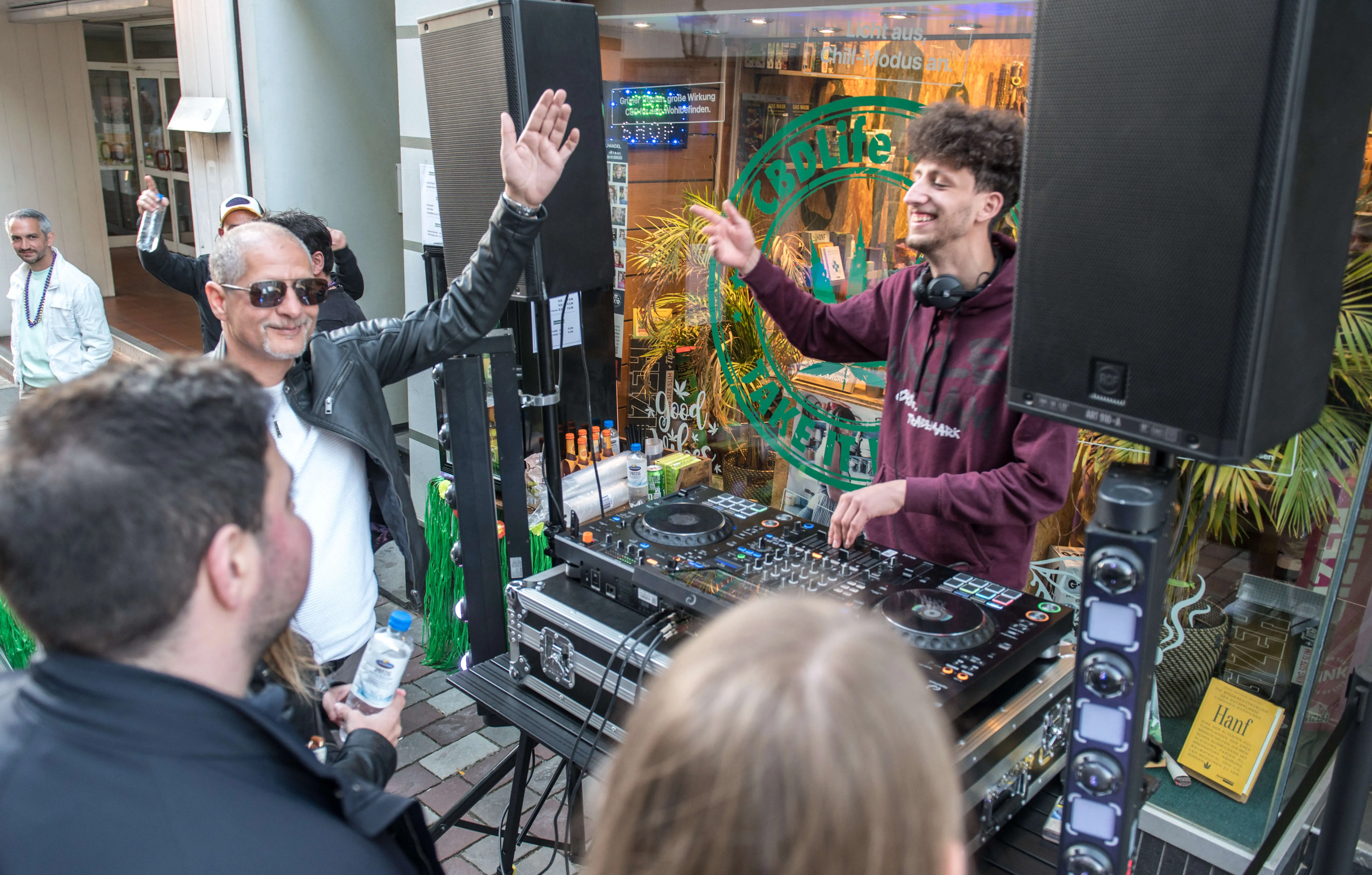 Zwei Männer stehen hinter einem DJ-Pult mit Plattenspielern und bedienen die Geräte. Im Hintergrund ist ein belebter Innenstadtbereich mit Geschäften und Menschen zu sehen.