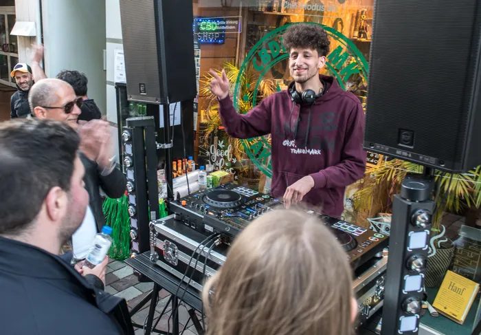 DJ spielt vor Publikum auf einer Bühne mit Equipment und Lautsprechern, im Hintergrund ein Geschäft mit Pflanzen und Schriftzügen.