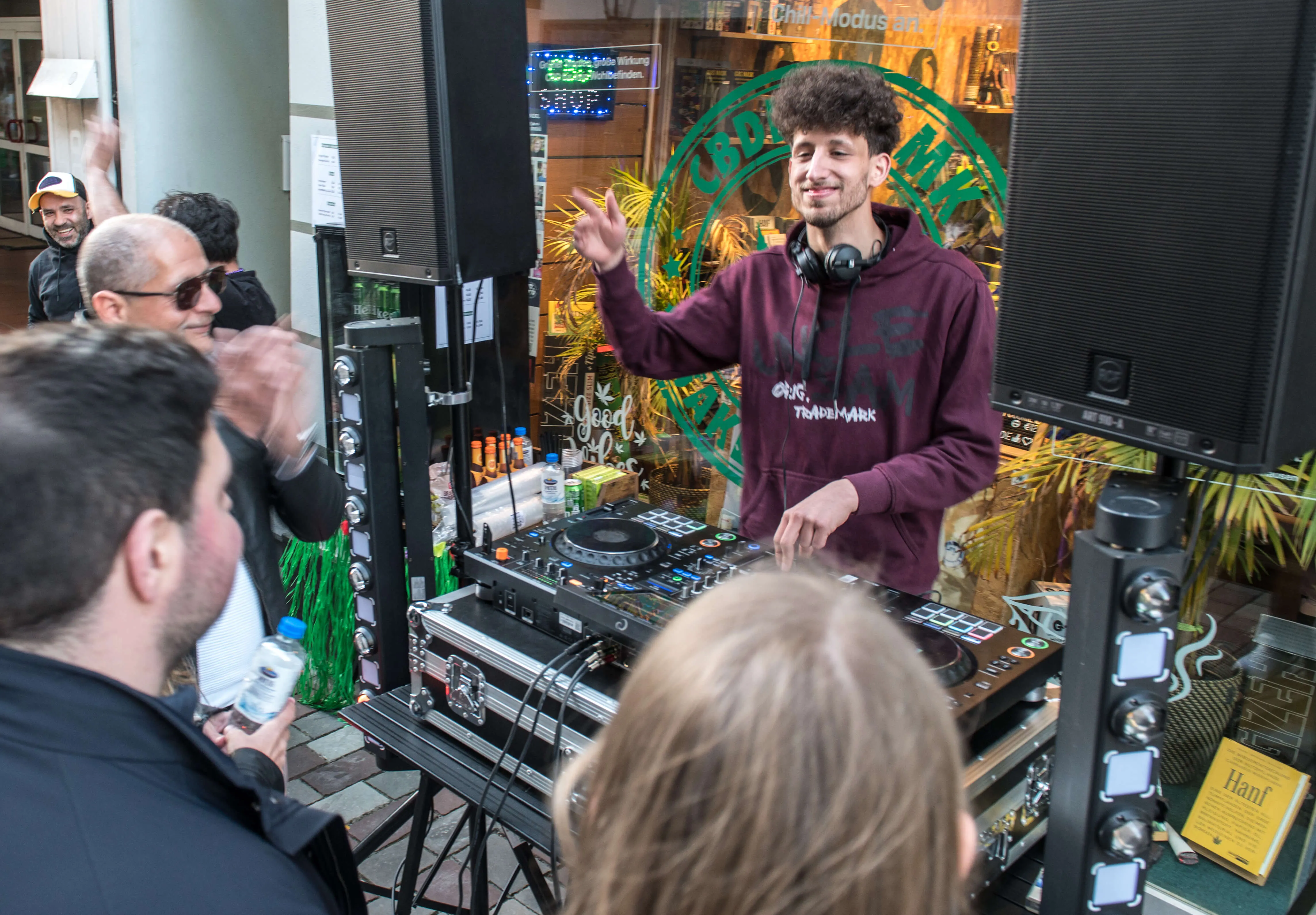 DJ spielt vor Publikum auf einer Bühne mit Equipment und Lautsprechern, im Hintergrund ein Geschäft mit Pflanzen und Schriftzügen.