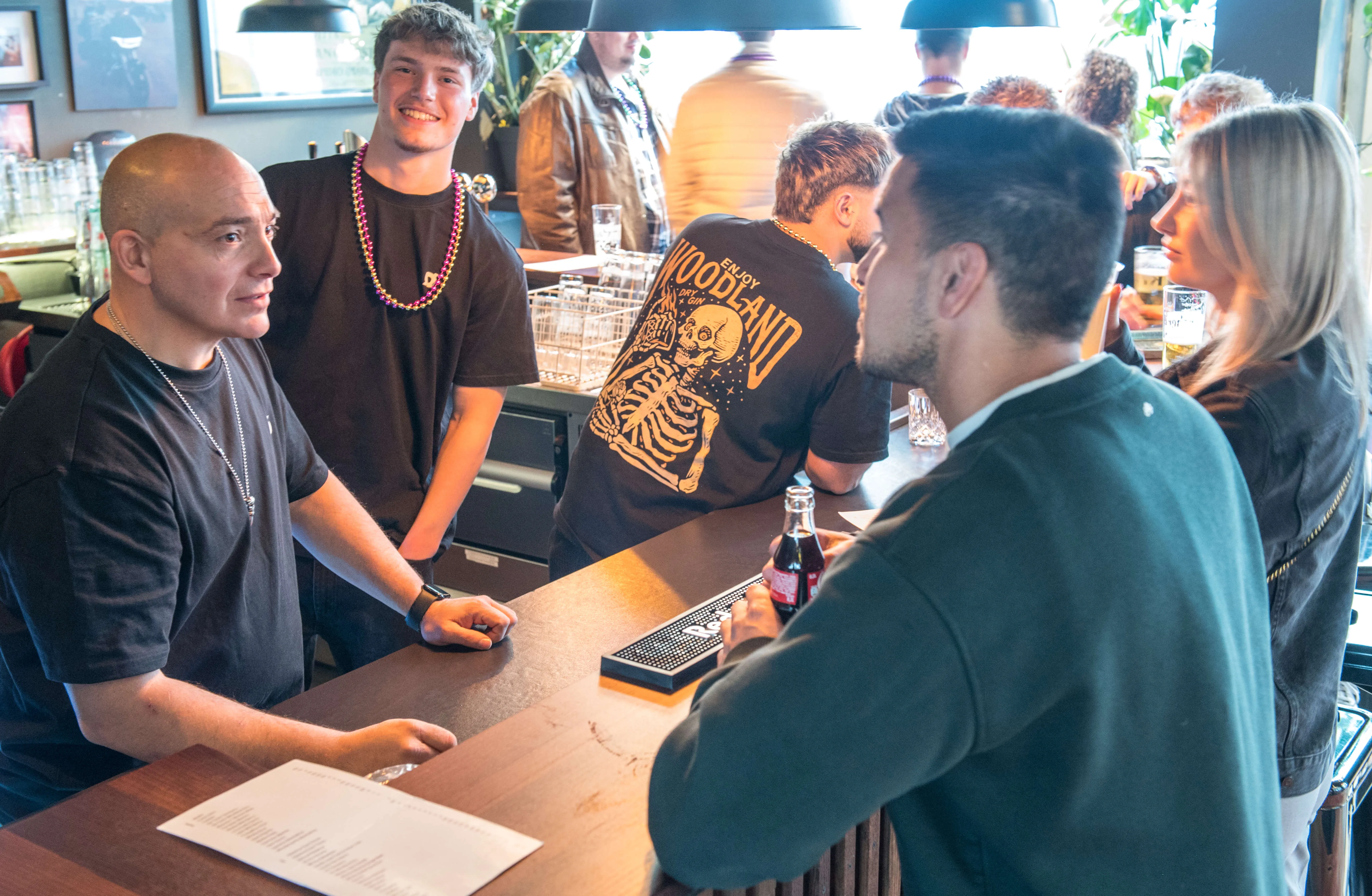 Mehrere Personen stehen an einer Bar, einige unterhalten sich und halten Getränke. Im Hintergrund sind weitere Menschen und ein belebter Innenraum zu sehen.