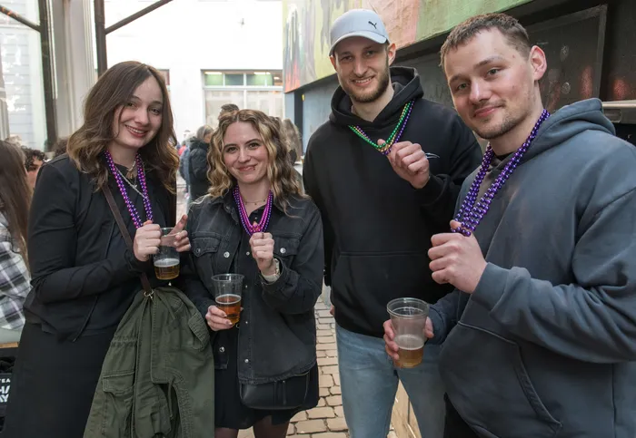 Vier junge Erwachsene mit Bier in der Hand und Karnevalsketten lächeln in die Kamera. Im Hintergrund sind weitere Menschen und eine belebte Straße zu sehen.