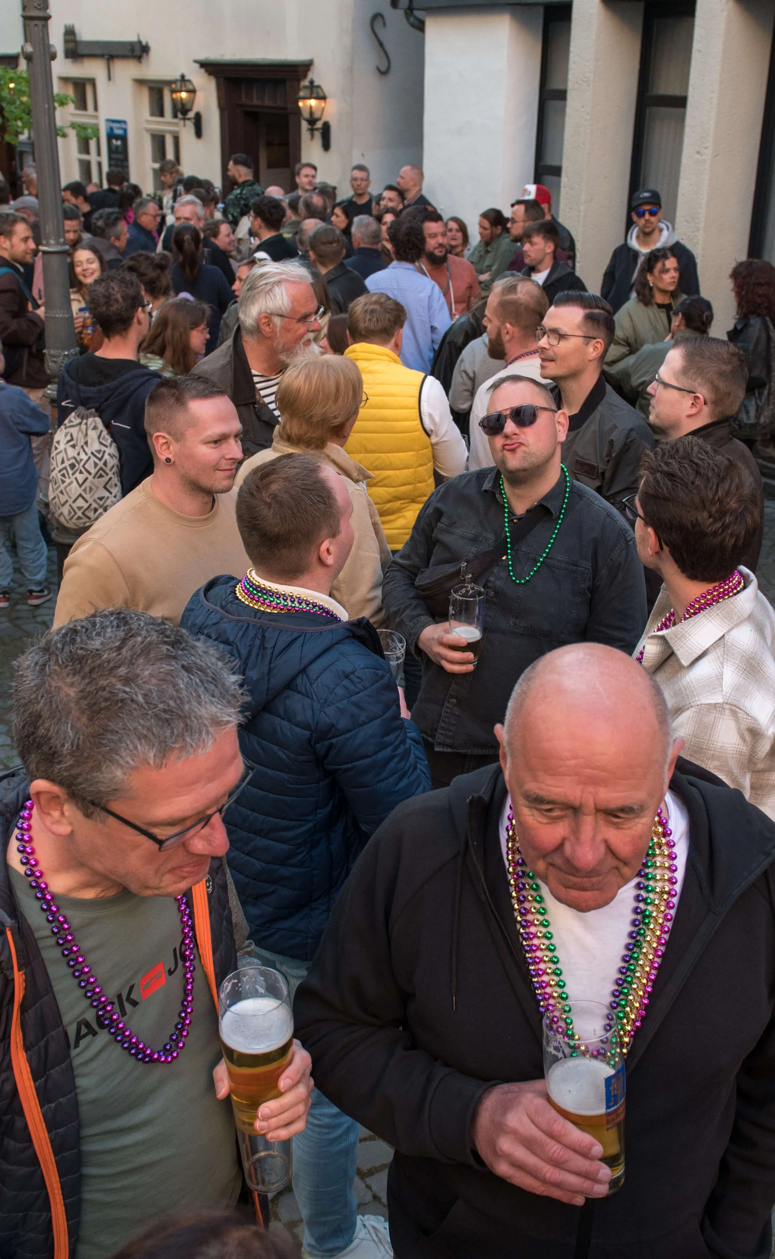 Menschen stehen dicht gedrängt vor einer Bar, viele halten Biergläser in der Hand und tragen bunte Perlenketten.
