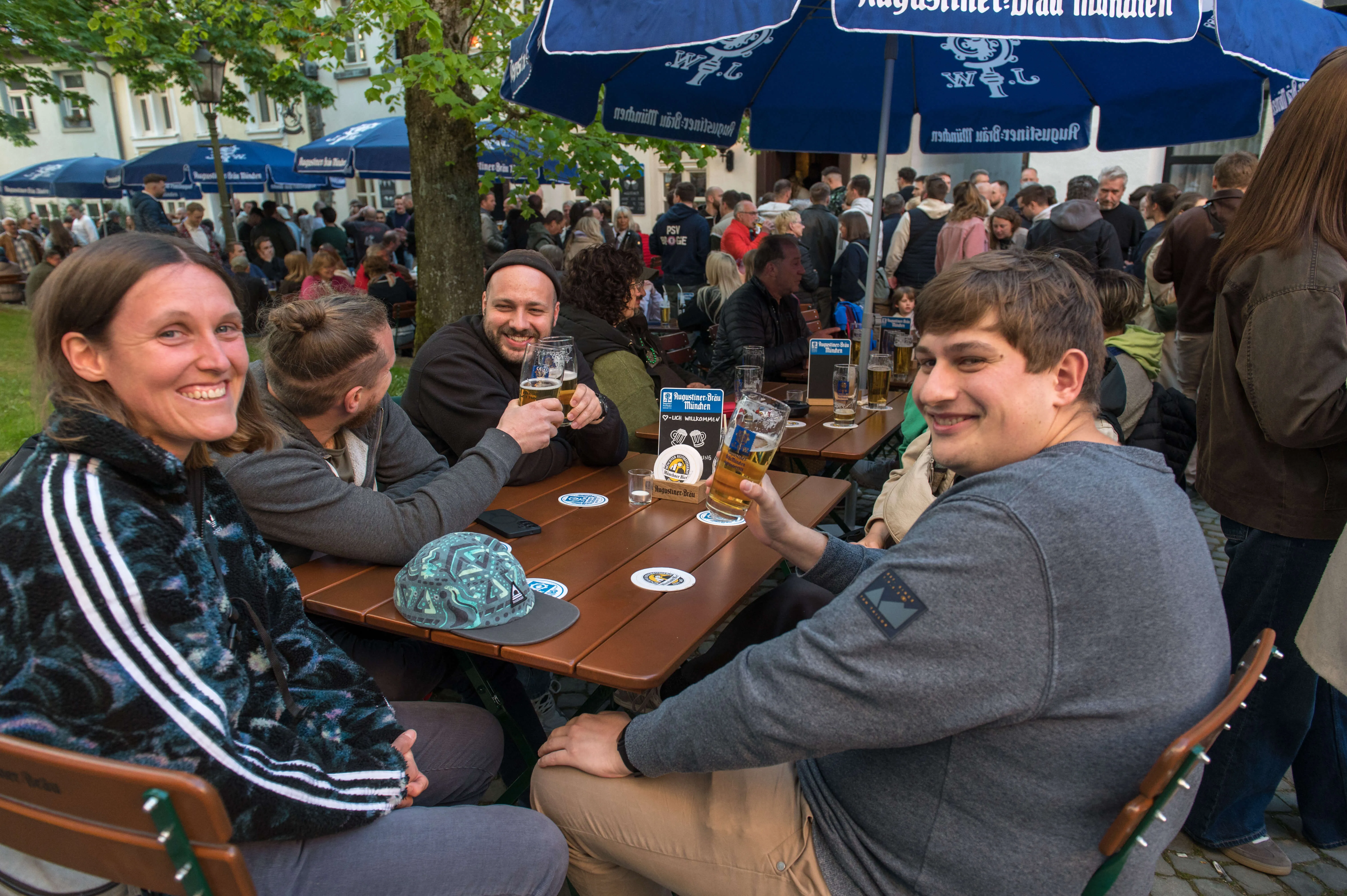 Drei Personen sitzen an einem Tisch im Freien, Biergläser davor. Im Hintergrund eine belebte Fußgängerzone mit weiteren Gästen und Schankstellen.