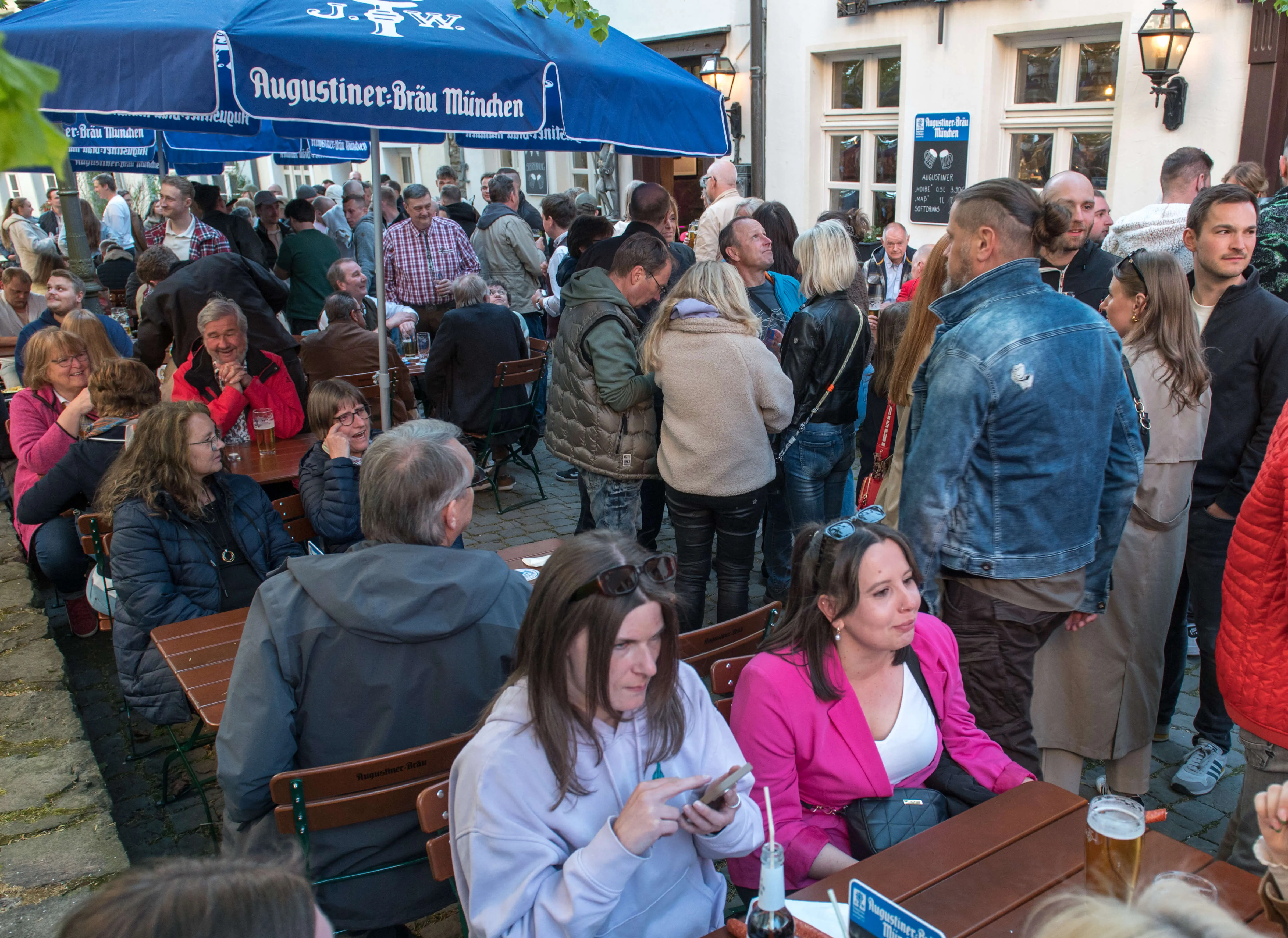 Menschen drängen sich auf einer belebten Straße vor einer Bar mit blauem Sonnenschirm, während sie Getränke genießen und sich unterhalten.