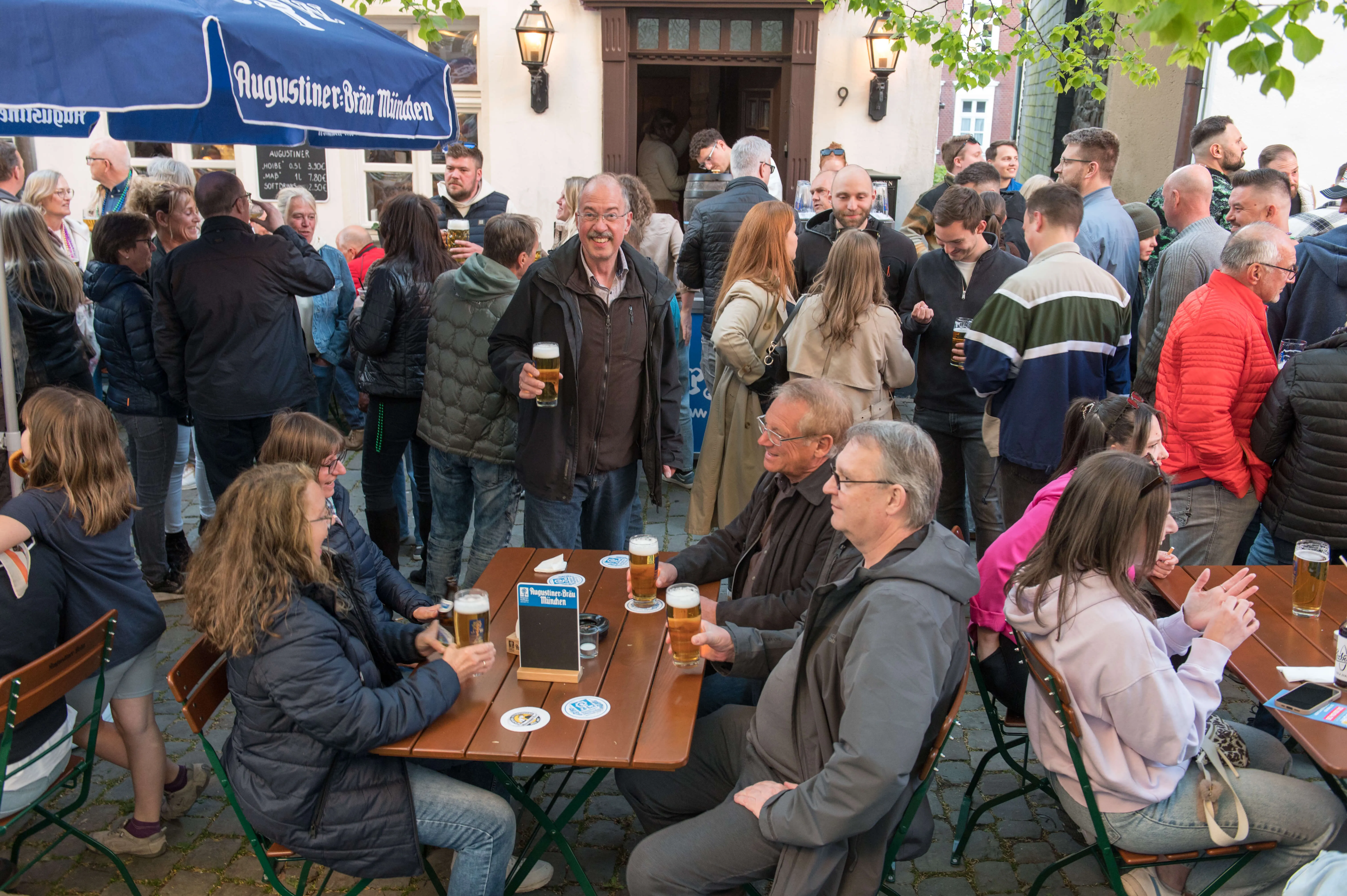 Menschen stehen und sitzen vor einer Kneipe im Freien, viele halten Bierkrüge in der Hand. Im Hintergrund ist ein Gebäude mit der Aufschrift „Augustiner-Bräu Lüdenscheid“ zu sehen.