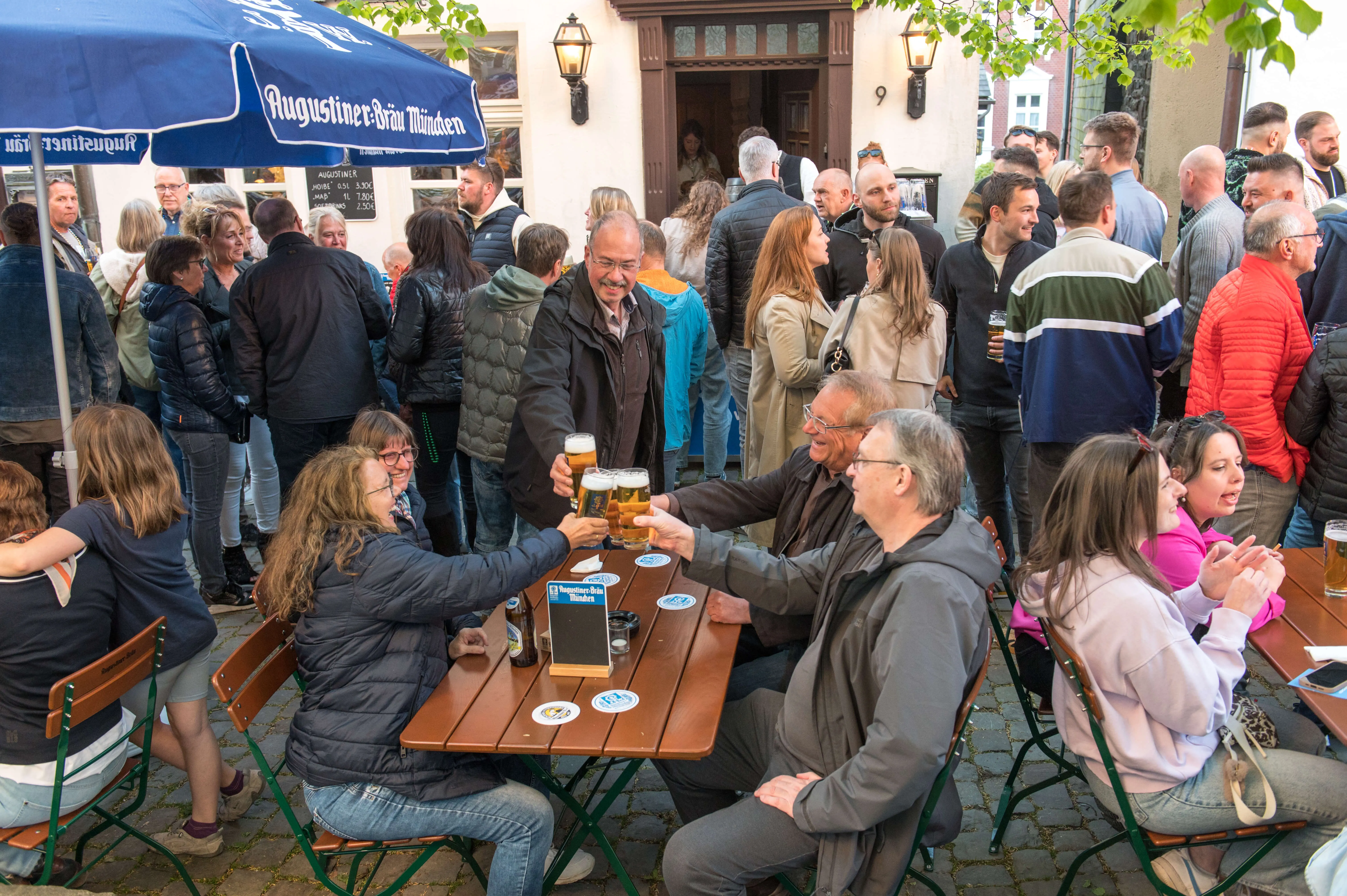 Menschen stehen und sitzen vor einer Bar im Freien, viele halten Bierkrüge in der Hand und stoßen an. Im Hintergrund ist eine belebte Straße mit weiteren Menschen zu sehen.