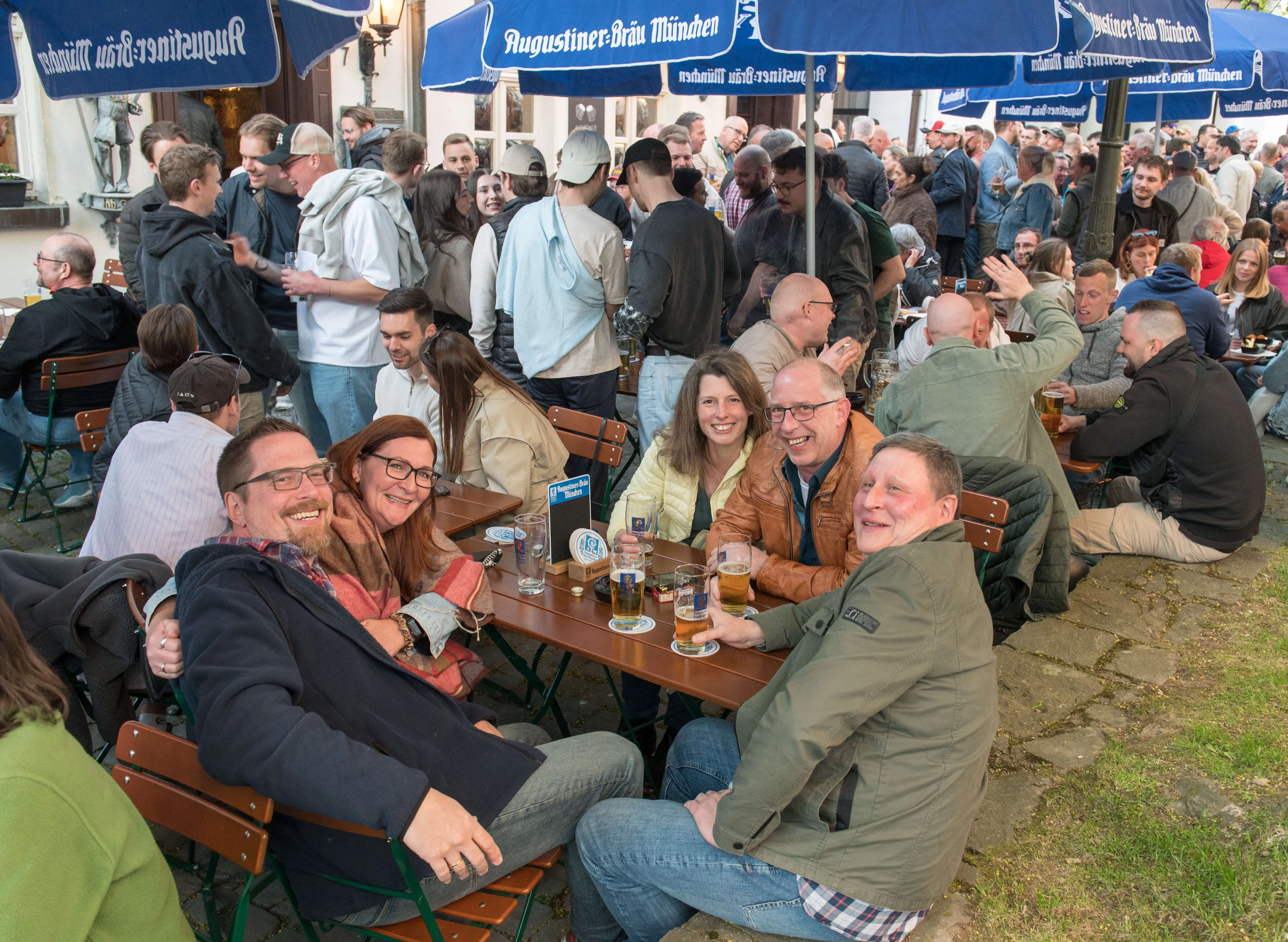 Menschen sitzen an Biertischgarnituren und genießen Getränke und Gespräche im Freien, umgeben von weiteren Besuchern und blau-weißen Sonnenschirmen während der Thekentour in Lüdenscheid.
