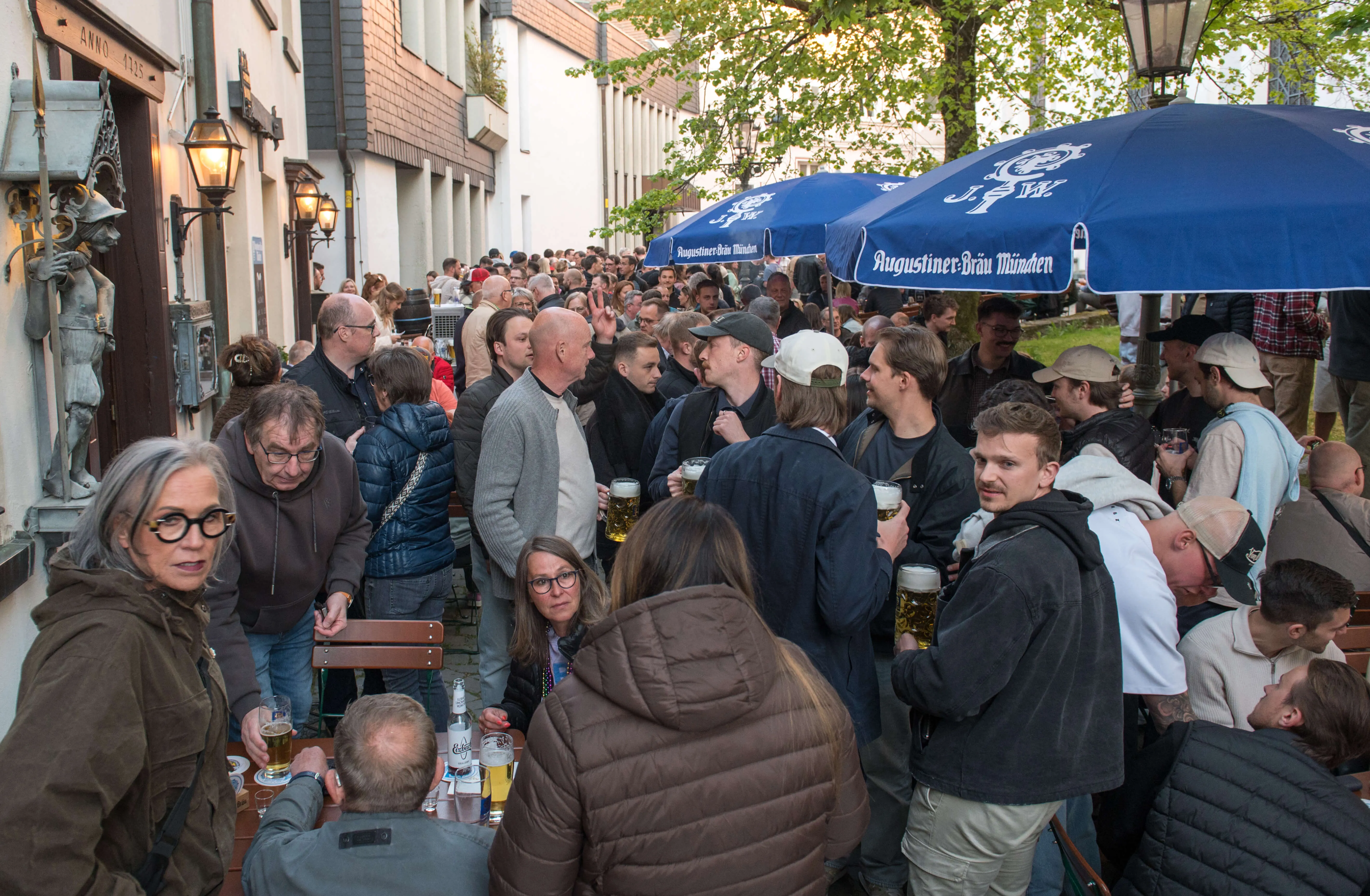 Menschen drängen sich auf einer belebten Straße vor Kneipen und Restaurants, viele halten Biergläser in der Hand. Blaue Sonnenschirme über den Tischen kennzeichnen die Außengastronomie.
