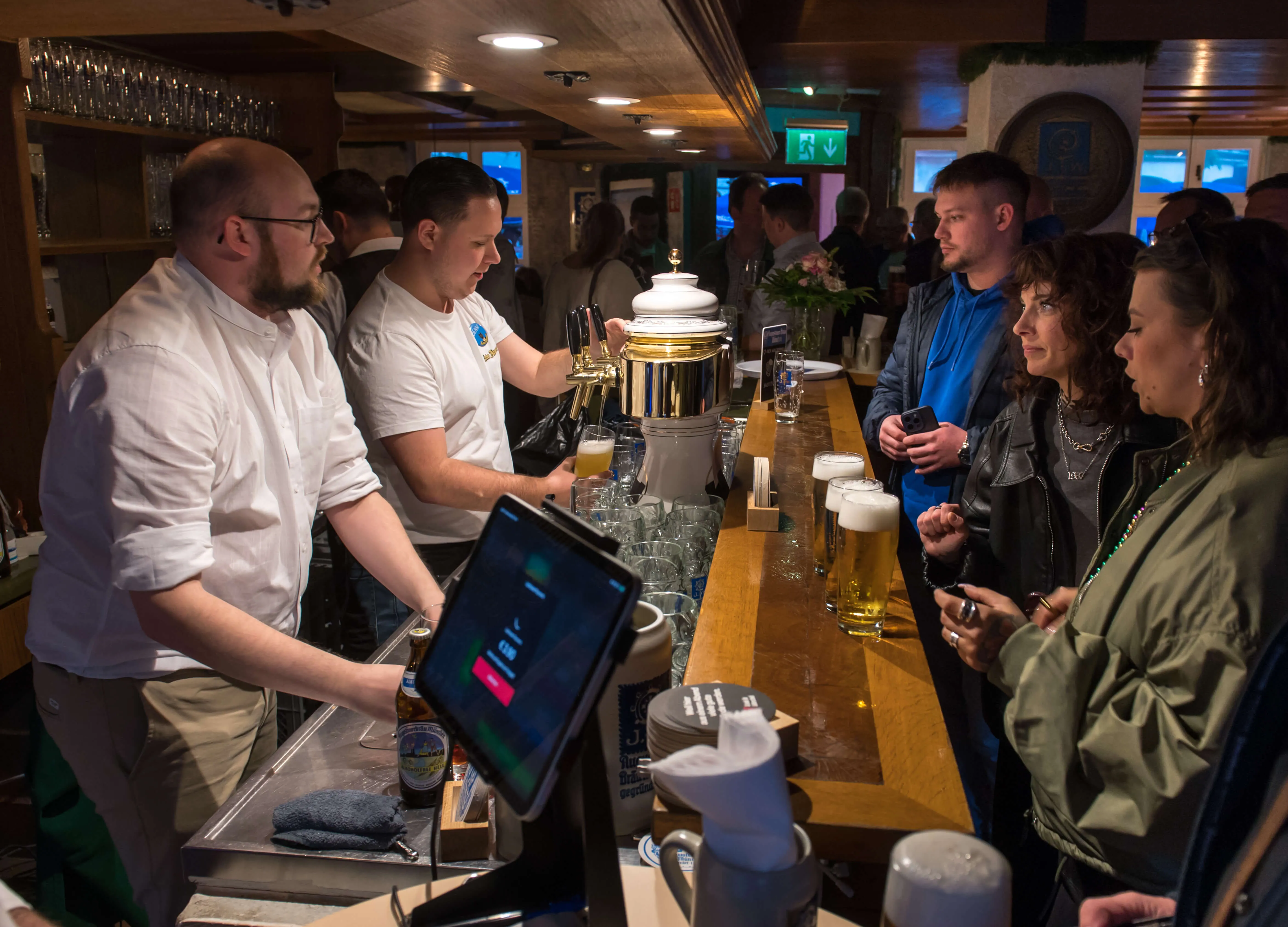 Zwei Barkeeper bedienen Gäste an einer Theke voller Biergläser während der Thekentour 2026 in Lüdenscheid. Im Hintergrund sind weitere Menschen zu sehen.