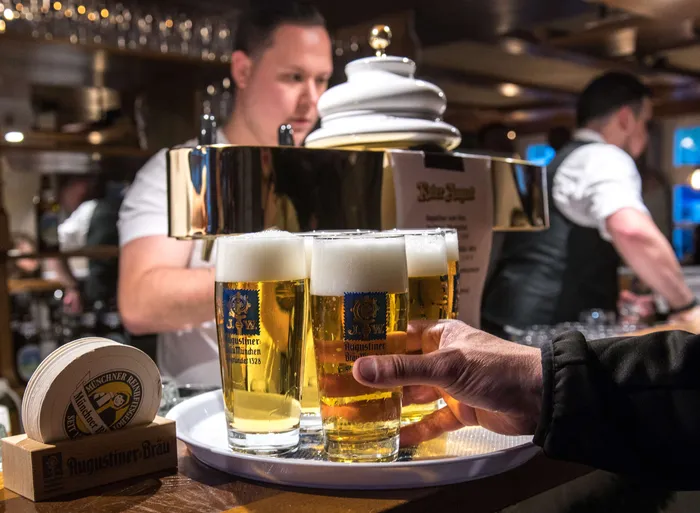 Ein Kellner serviert zwei volle Bierkrüge mit Augustiner-Bräu auf einem Tablett, im Hintergrund weitere Gäste in einer Bar.