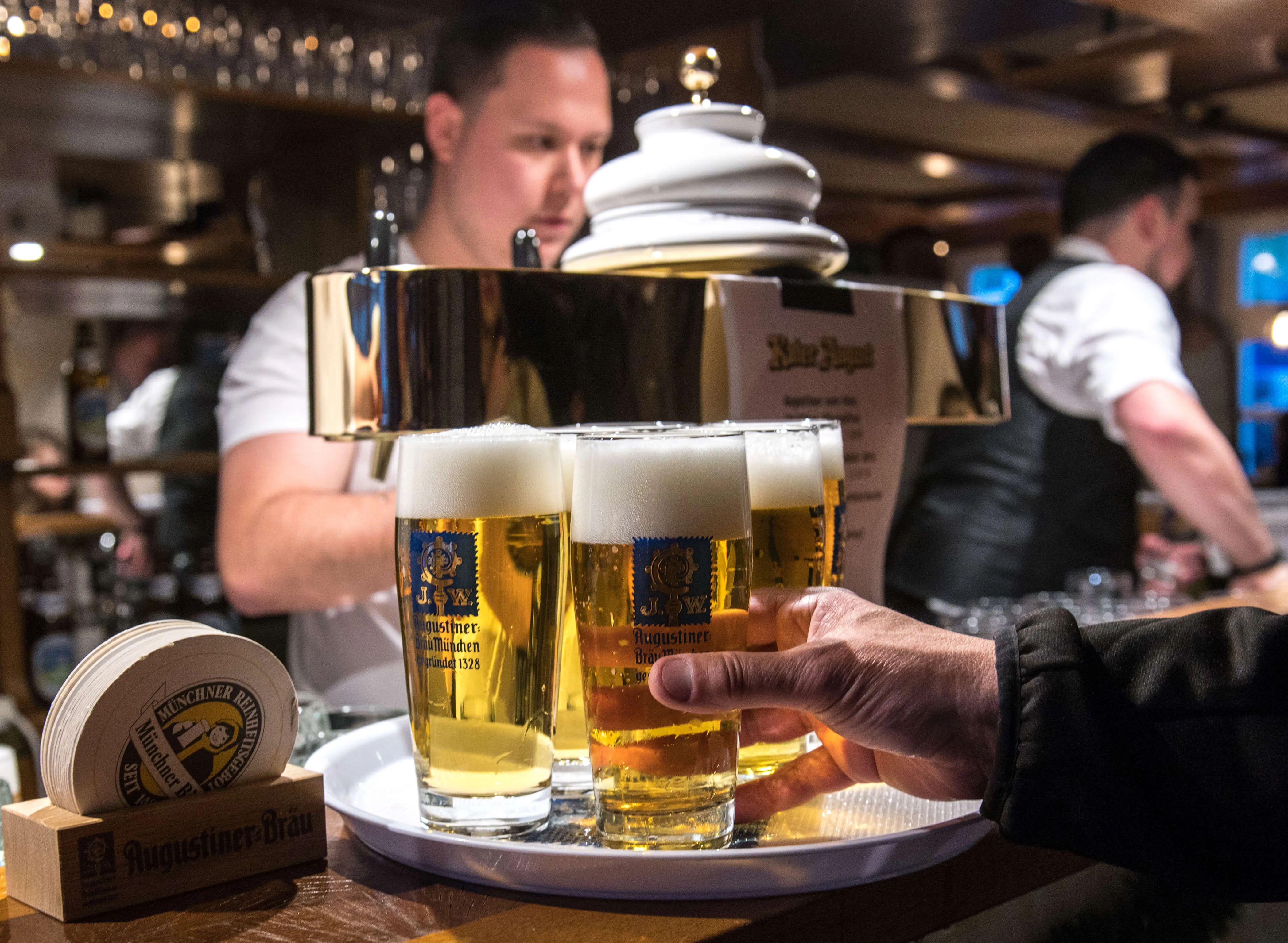 Ein Kellner serviert zwei volle Bierkrüge mit Augustiner-Bräu auf einem Tablett, im Hintergrund weitere Gäste in einer Bar.