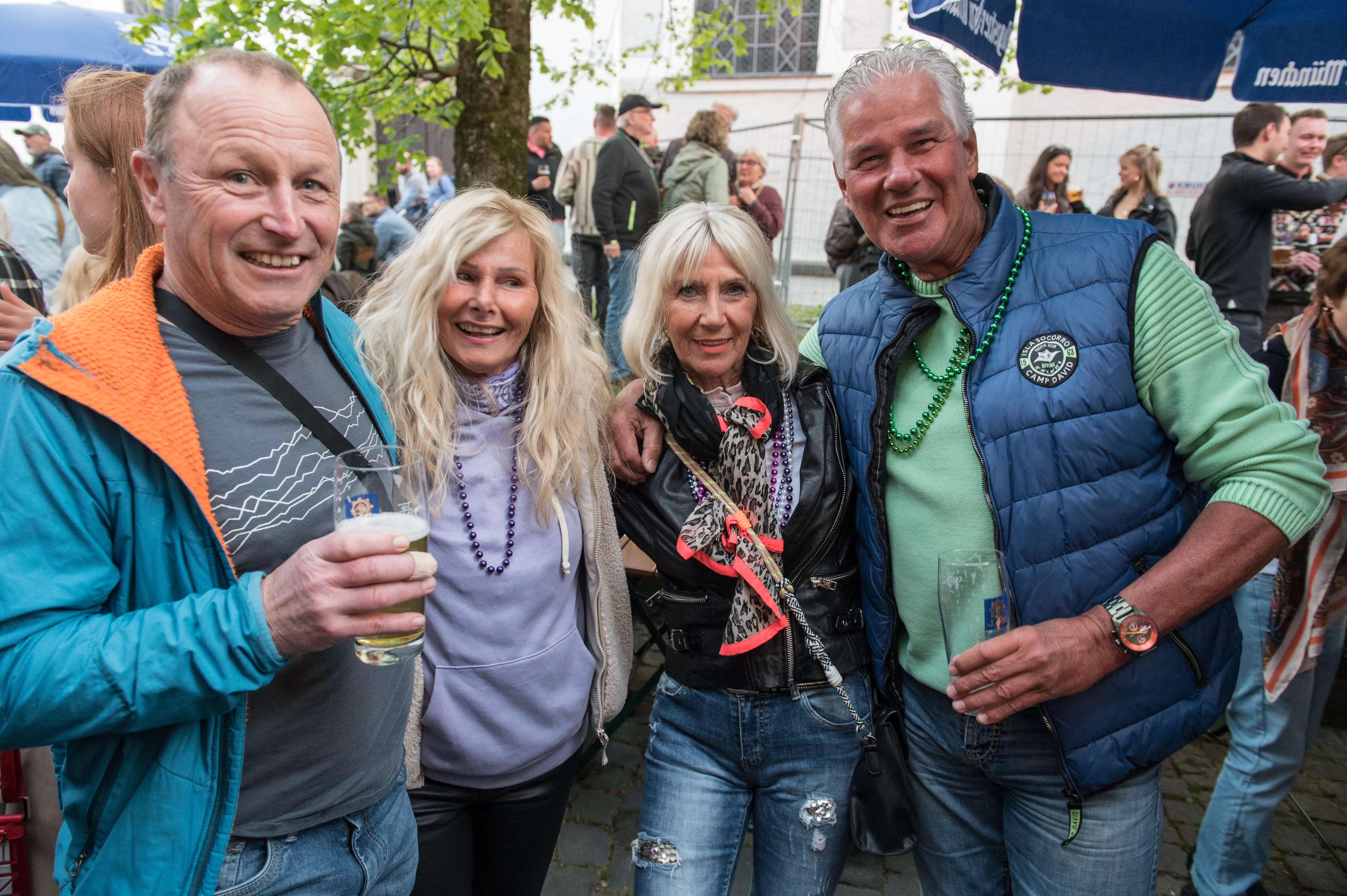 Vier lächelnde Menschen stehen eng zusammen, zwei Männer und zwei Frauen, inmitten einer belebten Menschenmenge bei einem Stadtfest.