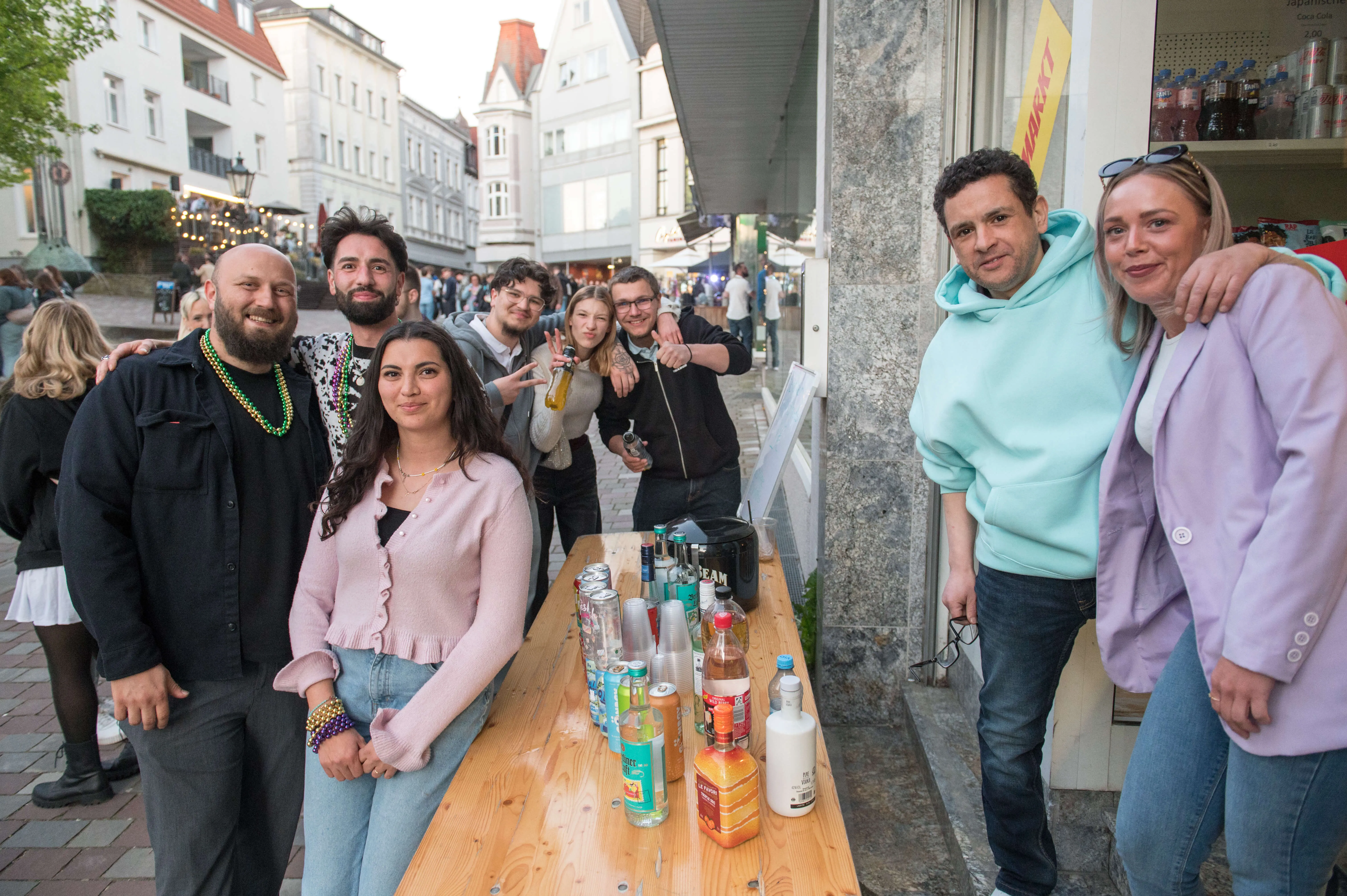 Eine Gruppe von jungen Erwachsenen steht lächelnd vor einem Tresen mit Getränken im Freien, im Hintergrund eine belebte Straße in Lüdenscheid während der Thekentour 2026.