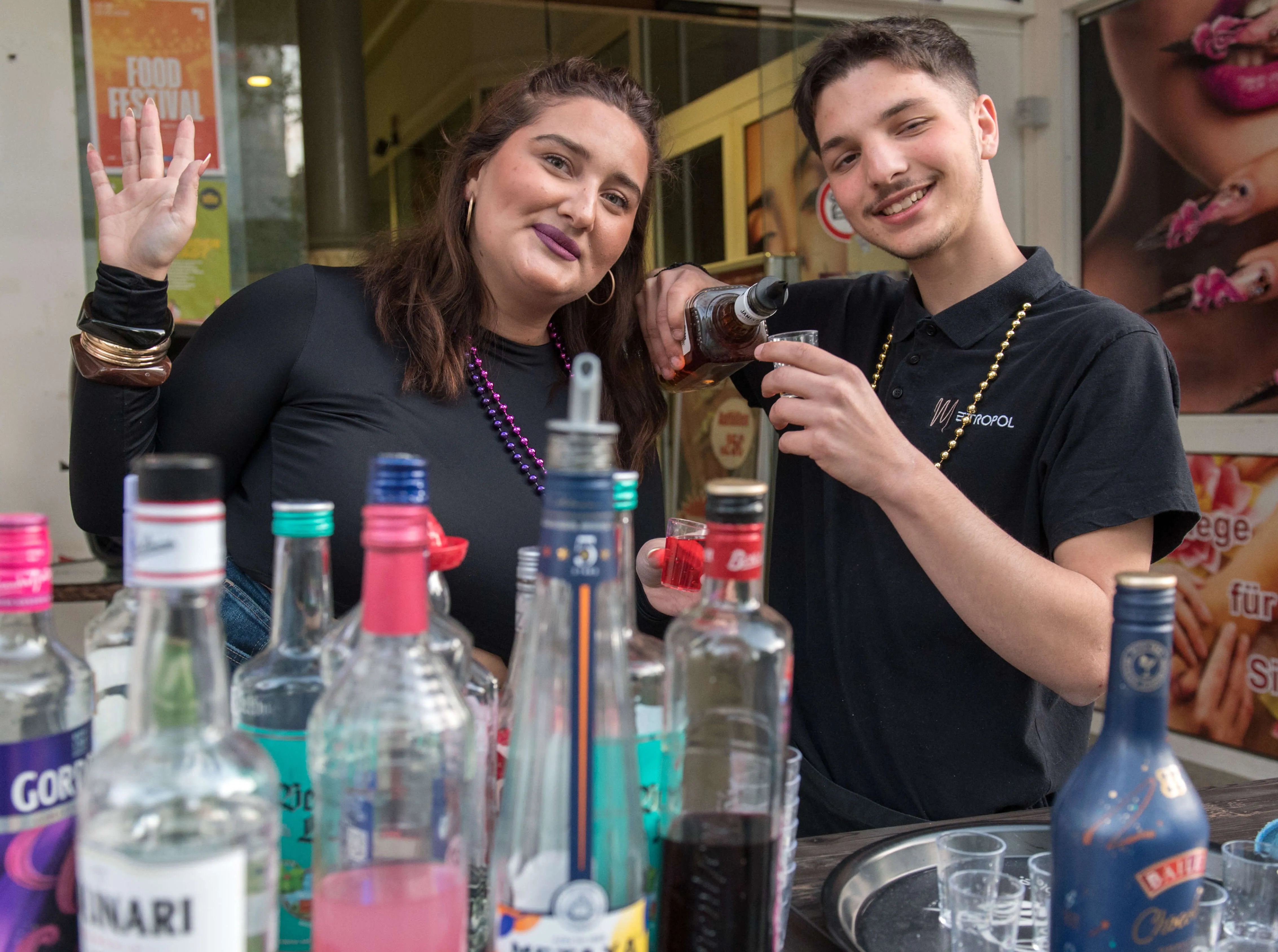 Zwei junge Menschen stehen hinter einer Bar mit verschiedenen Spirituosen und bereiten Getränke zu. Im Hintergrund ist ein Plakat mit der Aufschrift „Food Festival“ zu sehen.