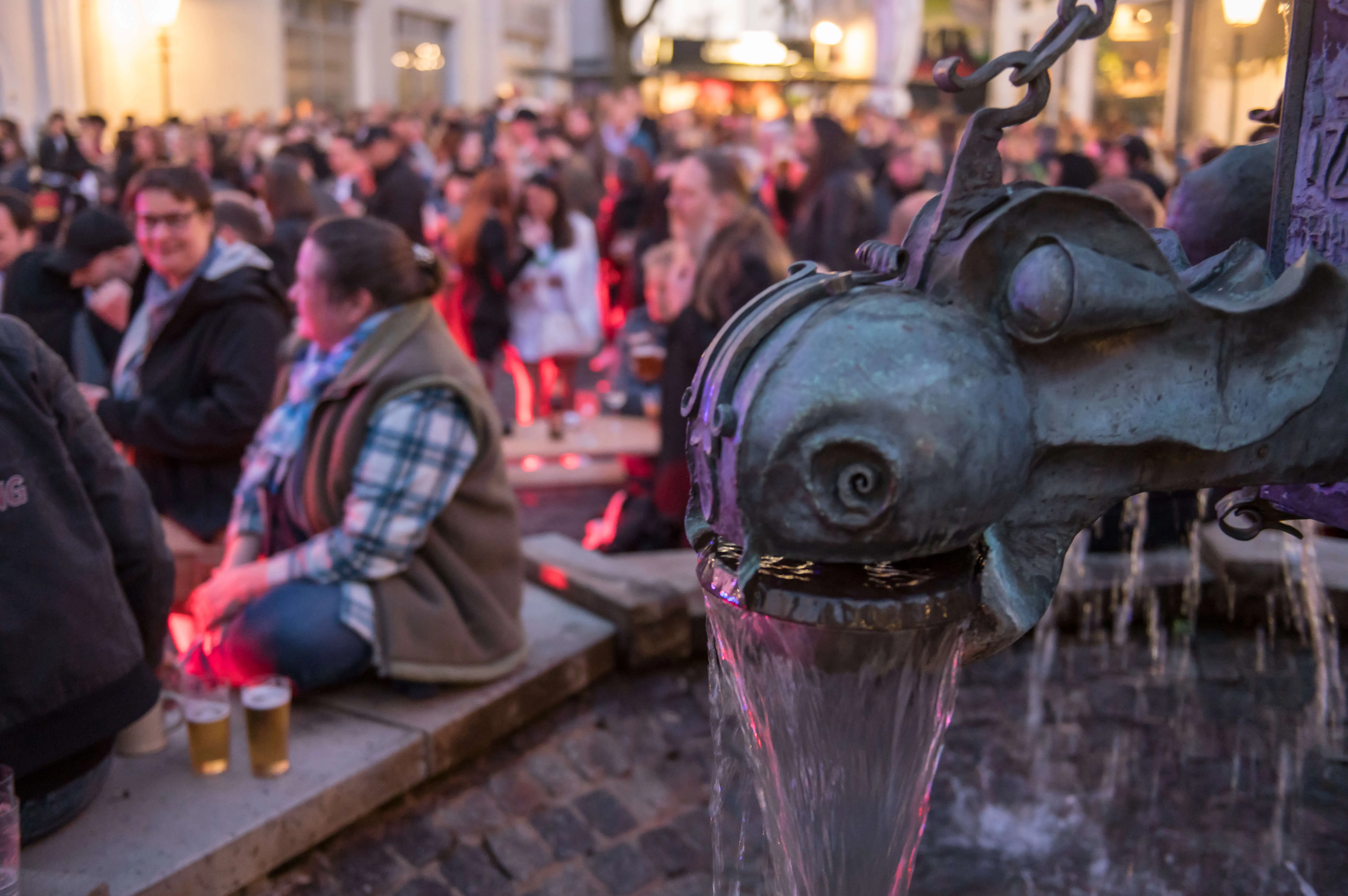 Menschen sitzen und stehen vor einer beleuchteten Bar, während Wasser aus dem Maul eines bronzenen Fisches in einem Brunnen fließt.