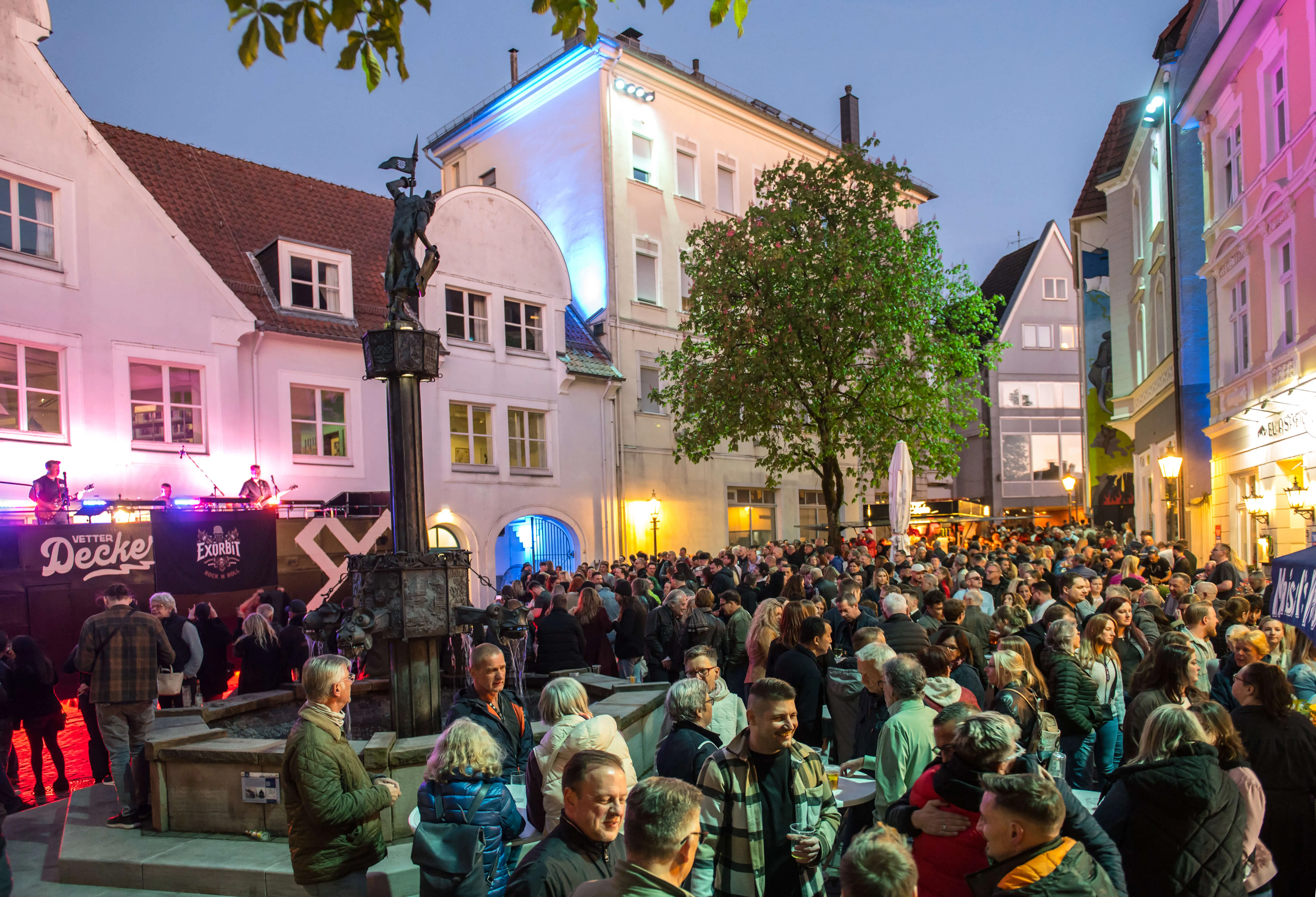 Große Menschenmenge auf einem Stadtplatz vor historischen Fachwerkhäusern während der Thekentour 2026 in Lüdenscheid. Im Hintergrund eine Bühne mit Musikern und ein beleuchteter Brunnen.