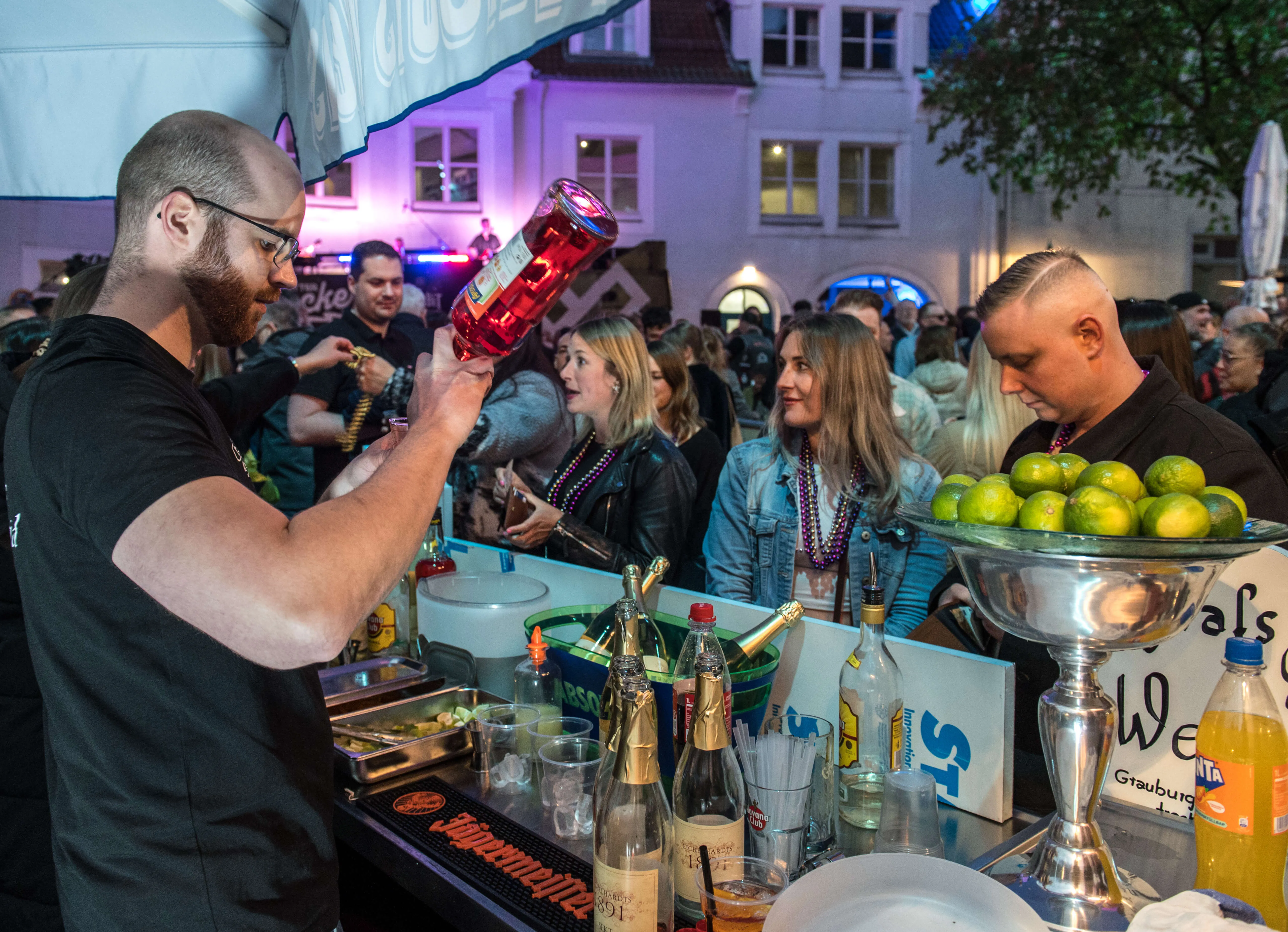 Zwei Barkeeper bereiten Getränke zu, während im Hintergrund Menschen auf einer belebten Straße feiern. Im Hintergrund sind Gebäude und Lichter zu sehen.