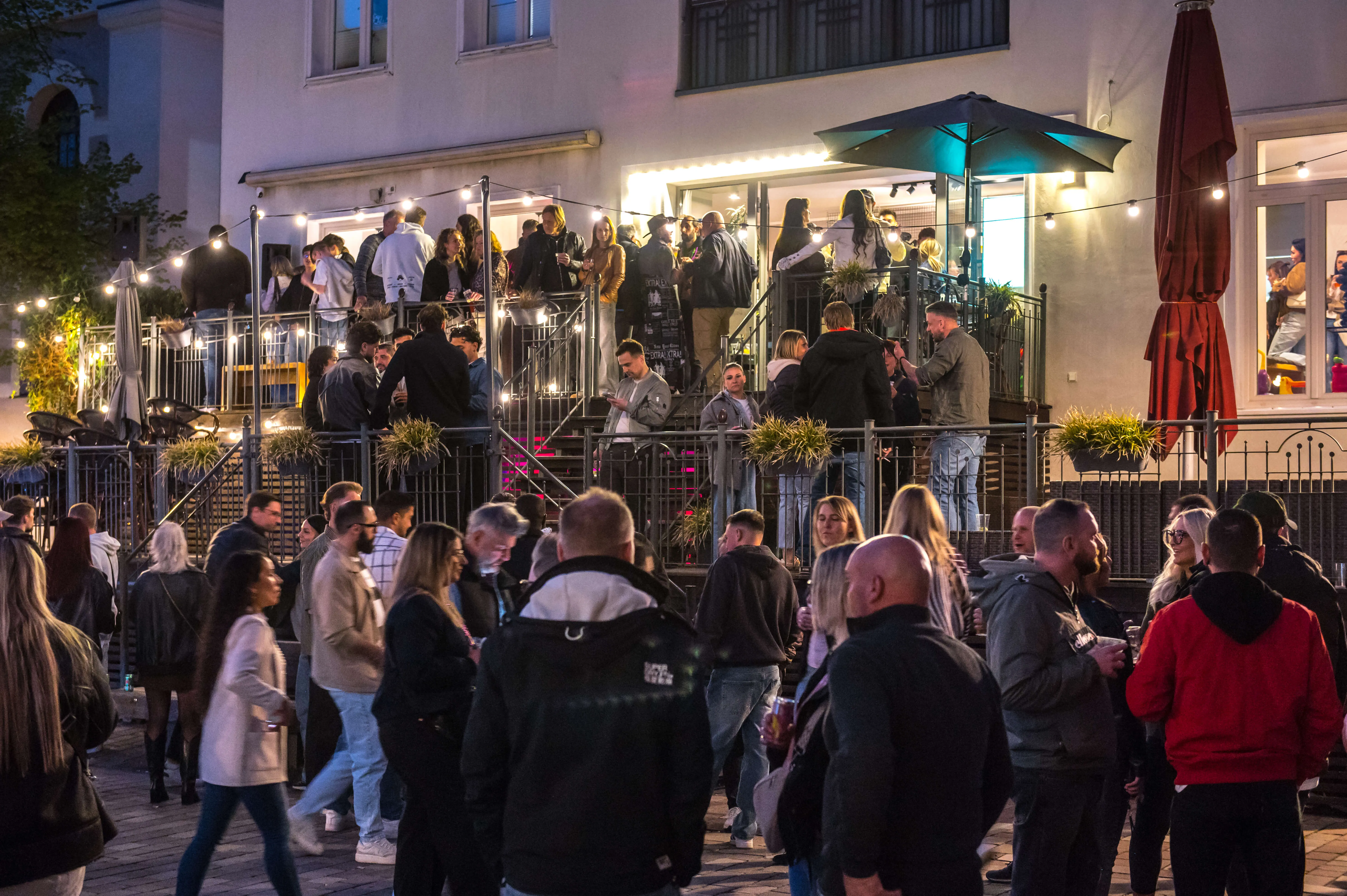 Menschen drängen sich auf einem belebten Platz vor einer Bar mit einer Treppe und einer Terrasse, die ebenfalls von Besuchern gefüllt ist.