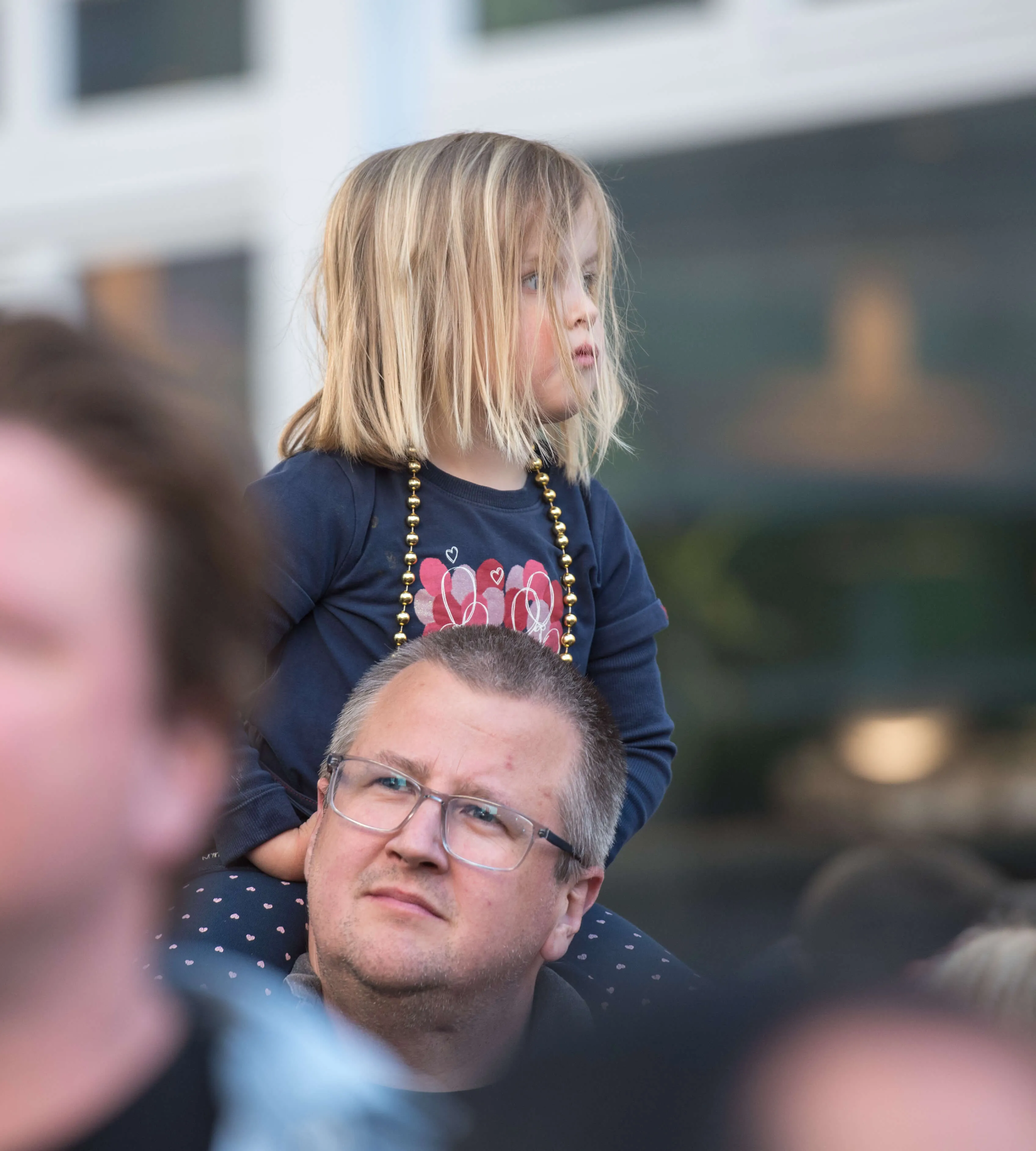Ein kleines Mädchen sitzt auf den Schultern eines Mannes und blickt zur Seite. Im Hintergrund sind verschwommene Menschen zu sehen.