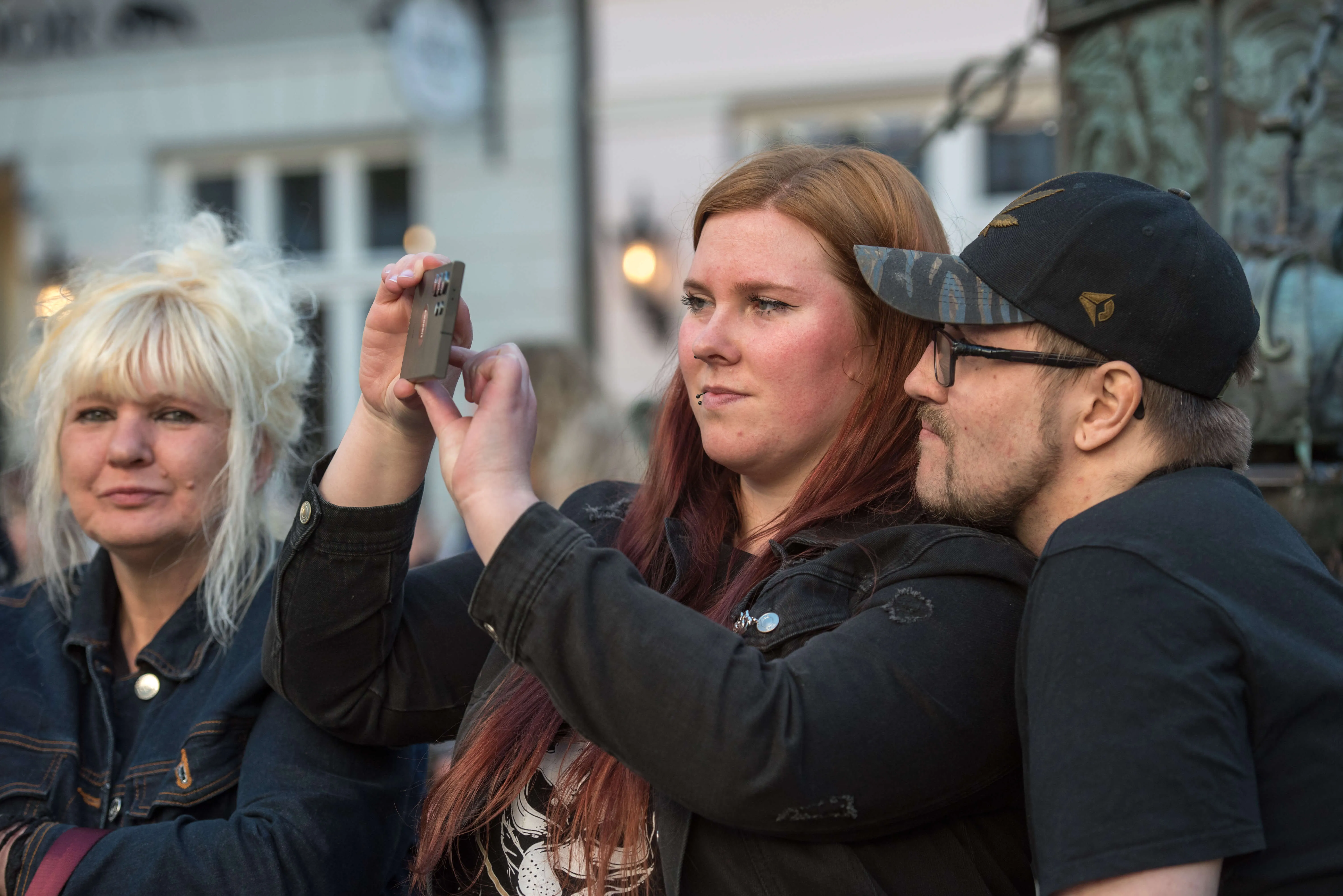 Drei junge Erwachsene fotografieren mit einem Smartphone während der Thekentour in Lüdenscheid. Im Hintergrund sind weitere Menschen und Gebäude zu sehen.