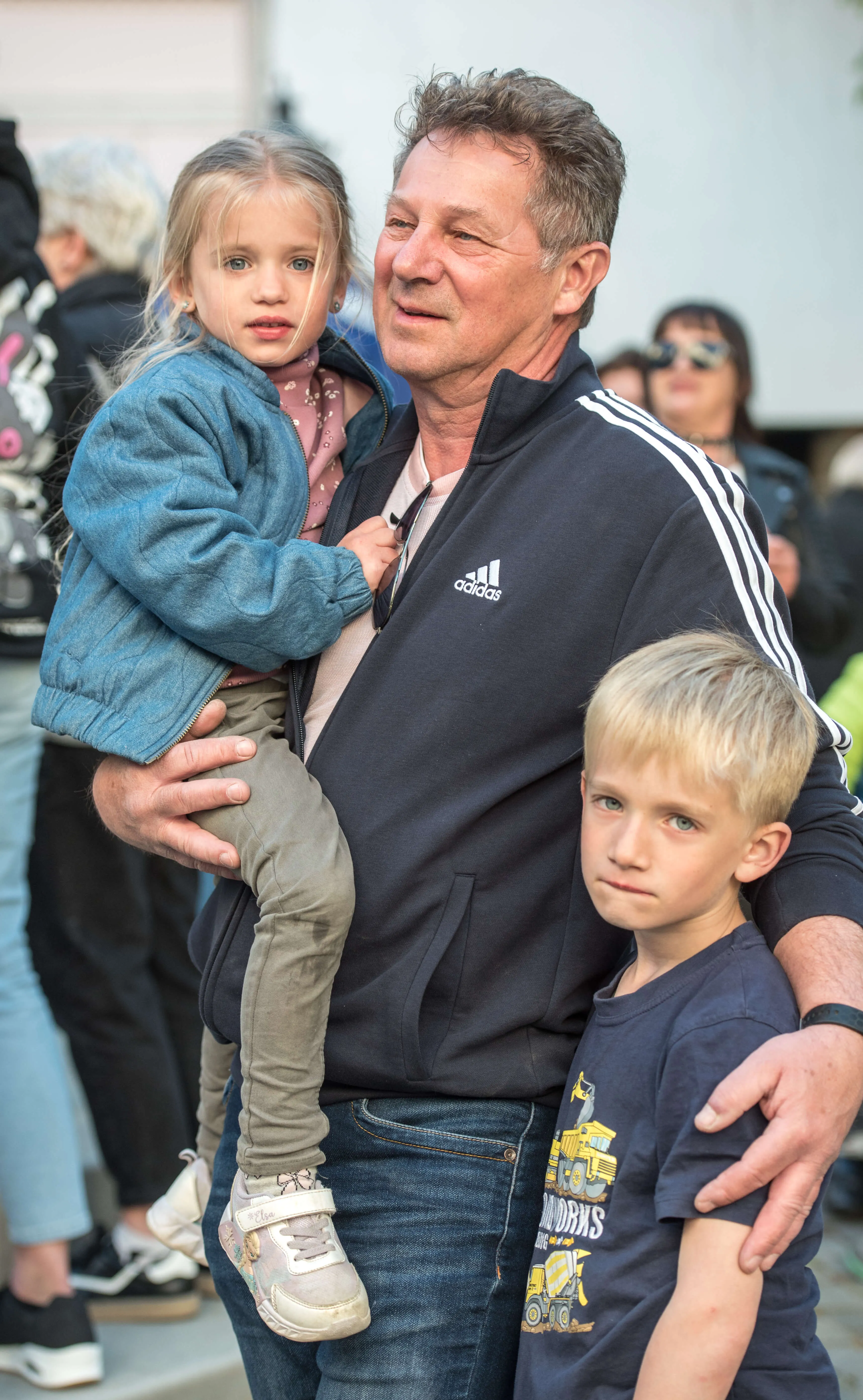 Ein Mann hält ein kleines Mädchen auf dem Arm, während er mit einem Jungen eng an sich gedrückt steht. Im Hintergrund sind weitere Menschen zu sehen.