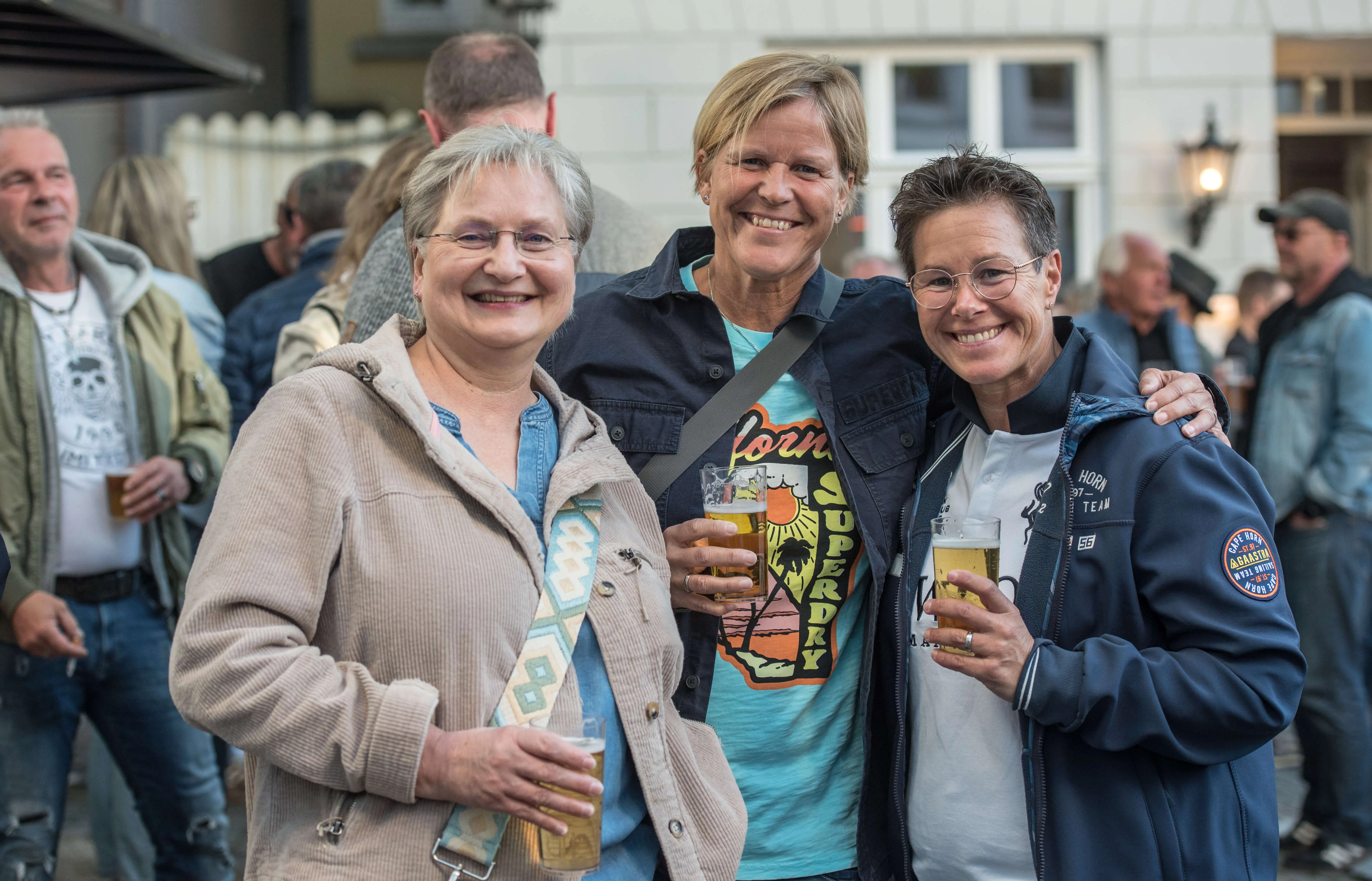 Drei lächelnde Frauen mittleren Alters stehen eng zusammen und halten jeweils ein Glas Bier in der Hand. Im Hintergrund sind weitere Menschen auf einer belebten Straße zu sehen.