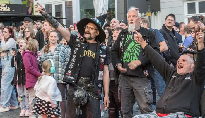 Mehrere Menschen feiern auf einer Straße, einige mit langen Haaren und in dunkler Kleidung, ein Mann trägt einen Hut und eine Weste mit Aufnähern.