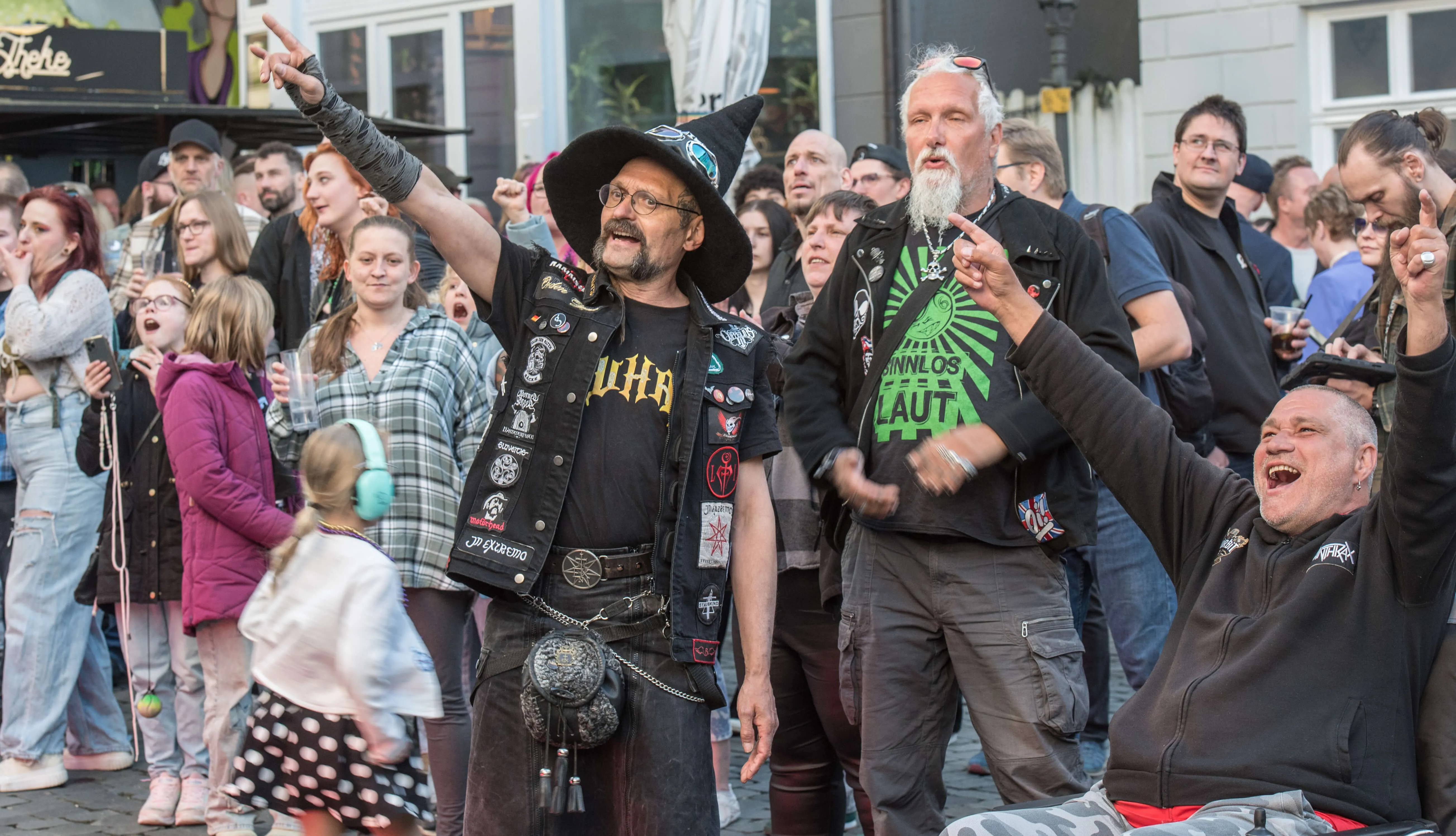 Mehrere Menschen feiern auf einer Straße, einige mit langen Haaren und in dunkler Kleidung, ein Mann trägt einen Hut und eine Weste mit Aufnähern.