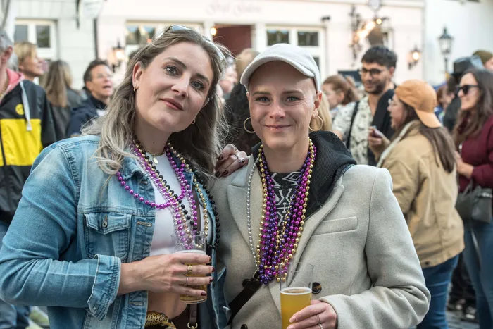 Zwei junge Frauen mit Perlenketten lächeln in die Kamera, jede hält ein Bierglas. Im Hintergrund ist eine Menschenmenge und ein Gebäude mit der Aufschrift „Zur Alten Mühle“ zu sehen.