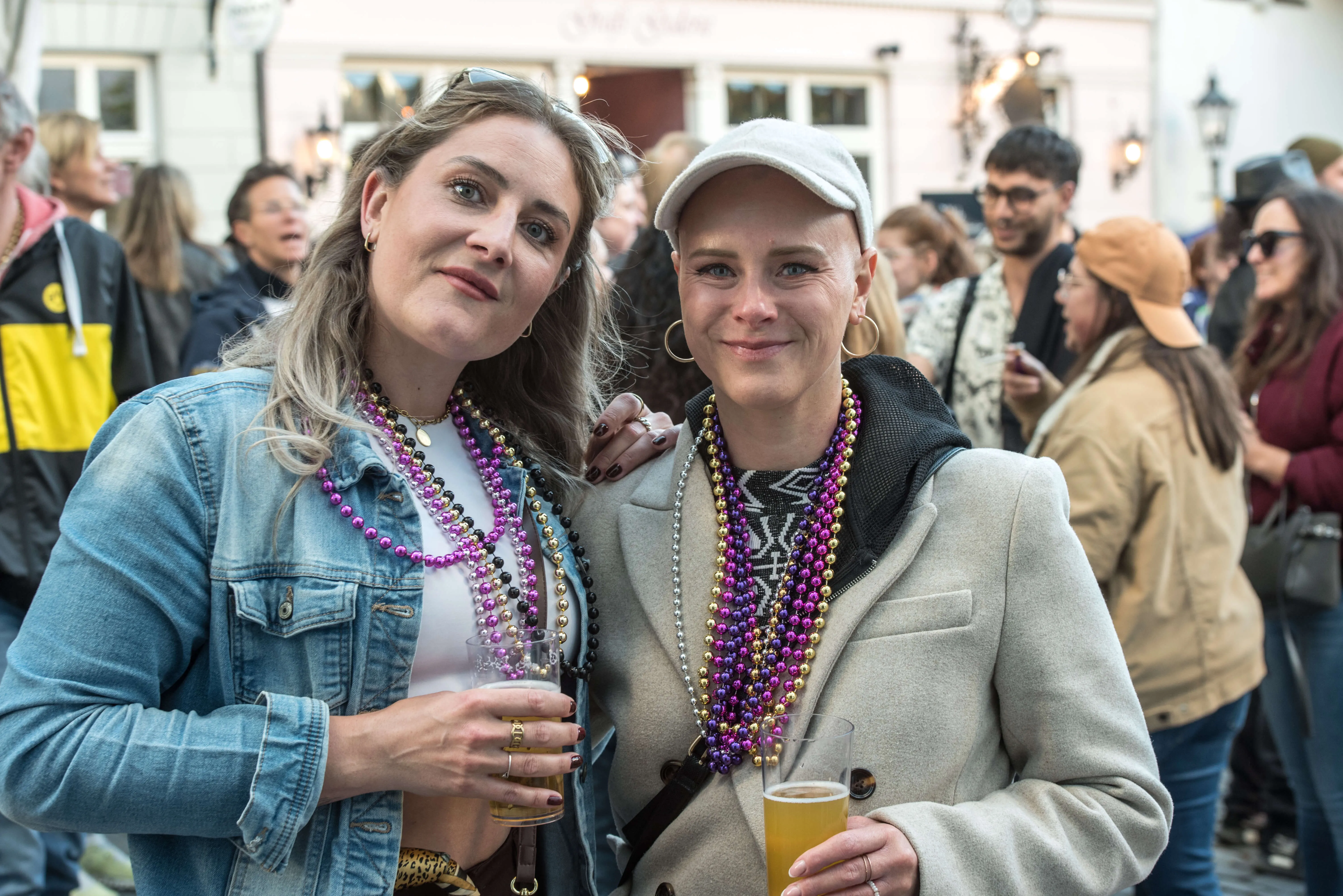 Zwei junge Frauen mit Perlenketten lächeln in die Kamera, jede hält ein Bierglas. Im Hintergrund ist eine Menschenmenge und ein Gebäude mit der Aufschrift „Zur Alten Mühle“ zu sehen.