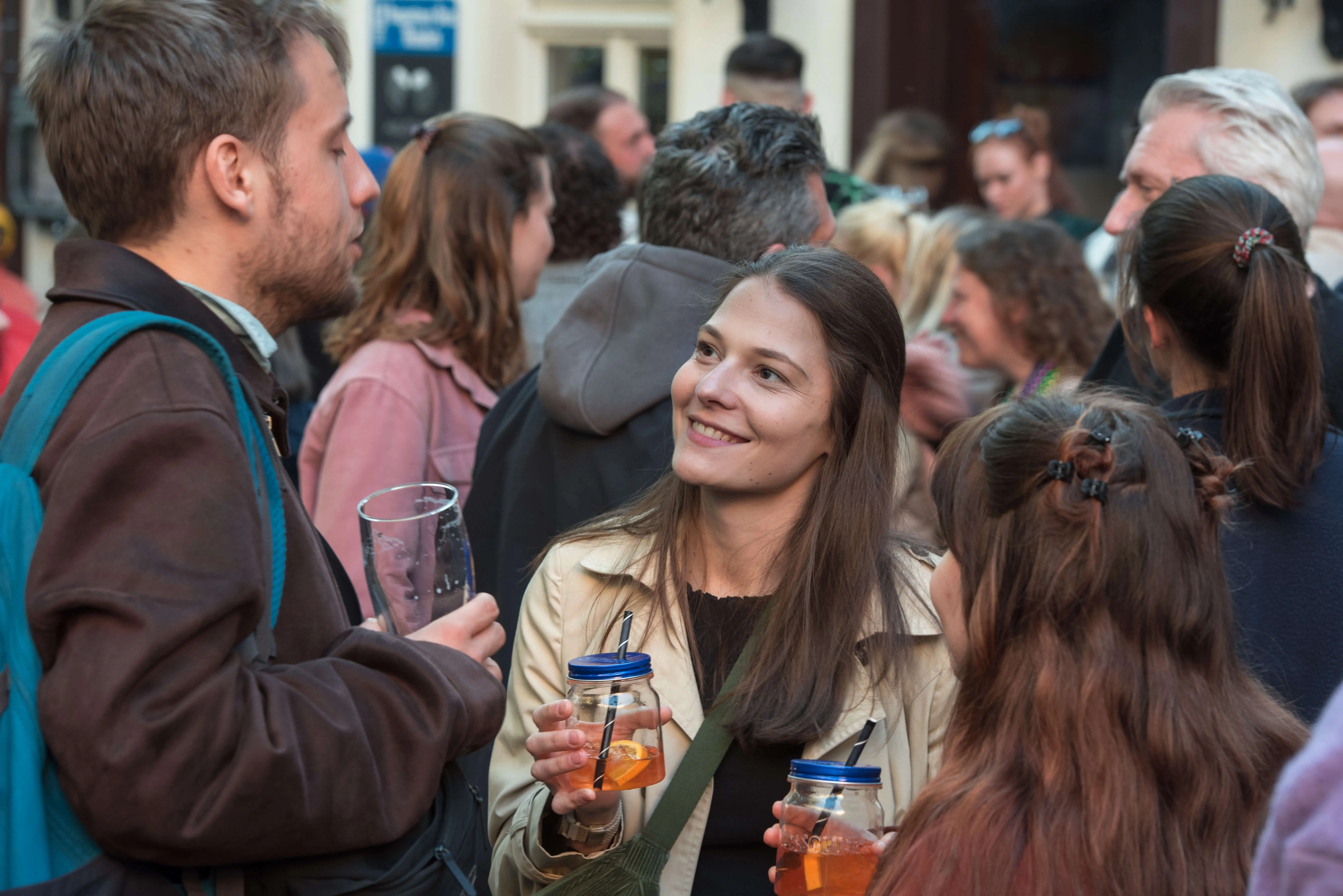 Eine junge Frau mit langen, braunen Haaren lächelt und blickt zur Seite, während sie ein Glas mit Getränk hält. Um sie herum sind viele Menschen zu sehen, die sich in einer belebten Kneipen- und Barlandschaft befinden.