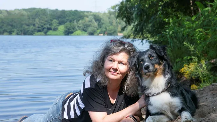 Therapeutin Anita Houben liegt am Ufer eines Sees und lächelt in die Kamera, neben ihr liegt ihr Australian Shepherd Lara.