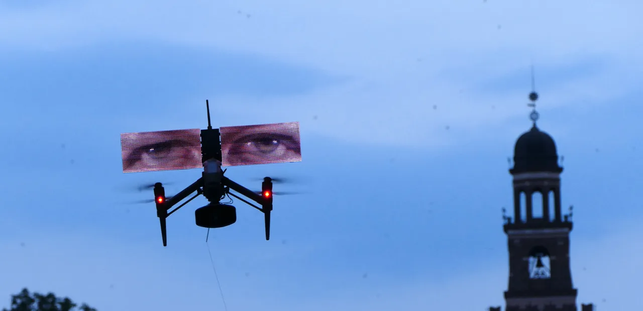 Eine Drohne mit einem LED-Bildschirm, der ein Paar Augen zeigt, fliegt vor einem Kirchturm am Abendhimmel.