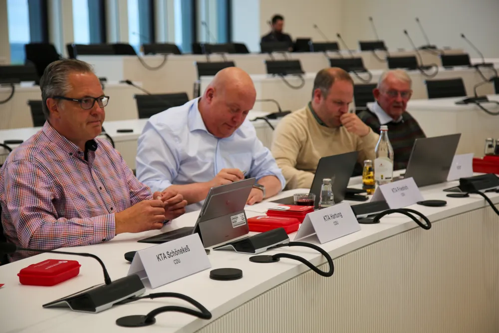 Vier Männer sitzen an einem Konferenztisch mit Laptops und Notizen, Namensschilder vor ihnen identifizieren sie als Ausschussmitglieder des Märkischen Kreises.