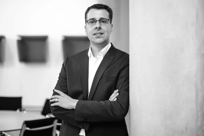 Thomas Schielke steht mit verschränkten Armen in einem dunklen Anzug und Brille in einem Schwarz-Weiß-Porträt.